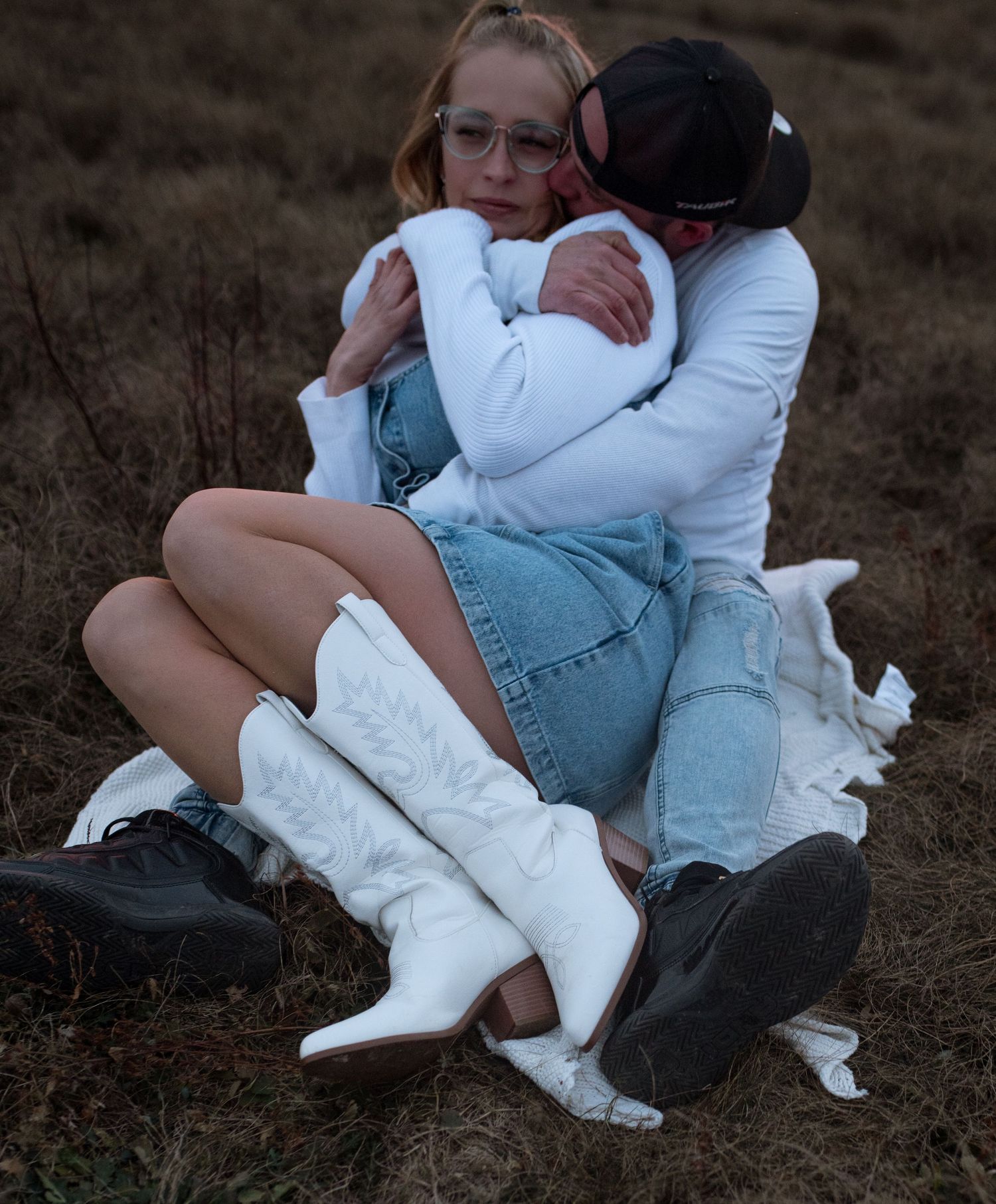 Couple embracing outdoors, woman in white cowboy boots and denim skirt sitting with partner in field.