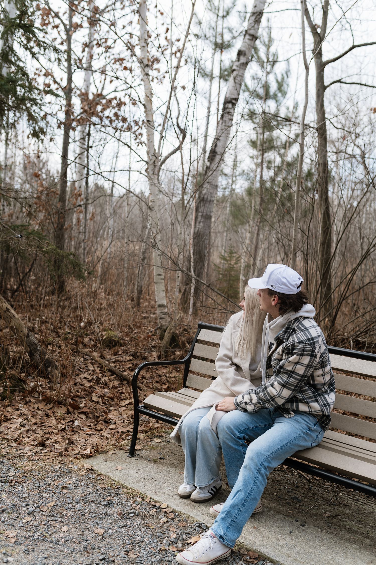 Two people sitting close together on a park bench surrounded by bare autumn trees on a wooded trail.