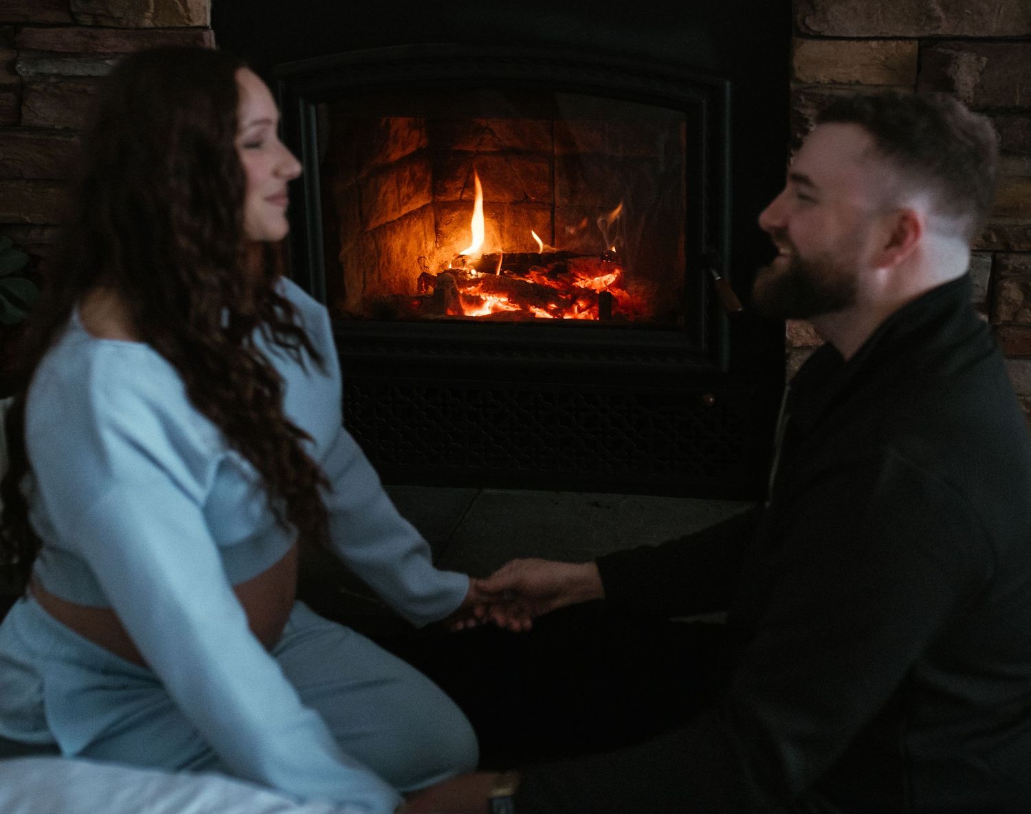 Pregnant woman and man holding hands facing each other in front of a warm glowing stone fireplace.