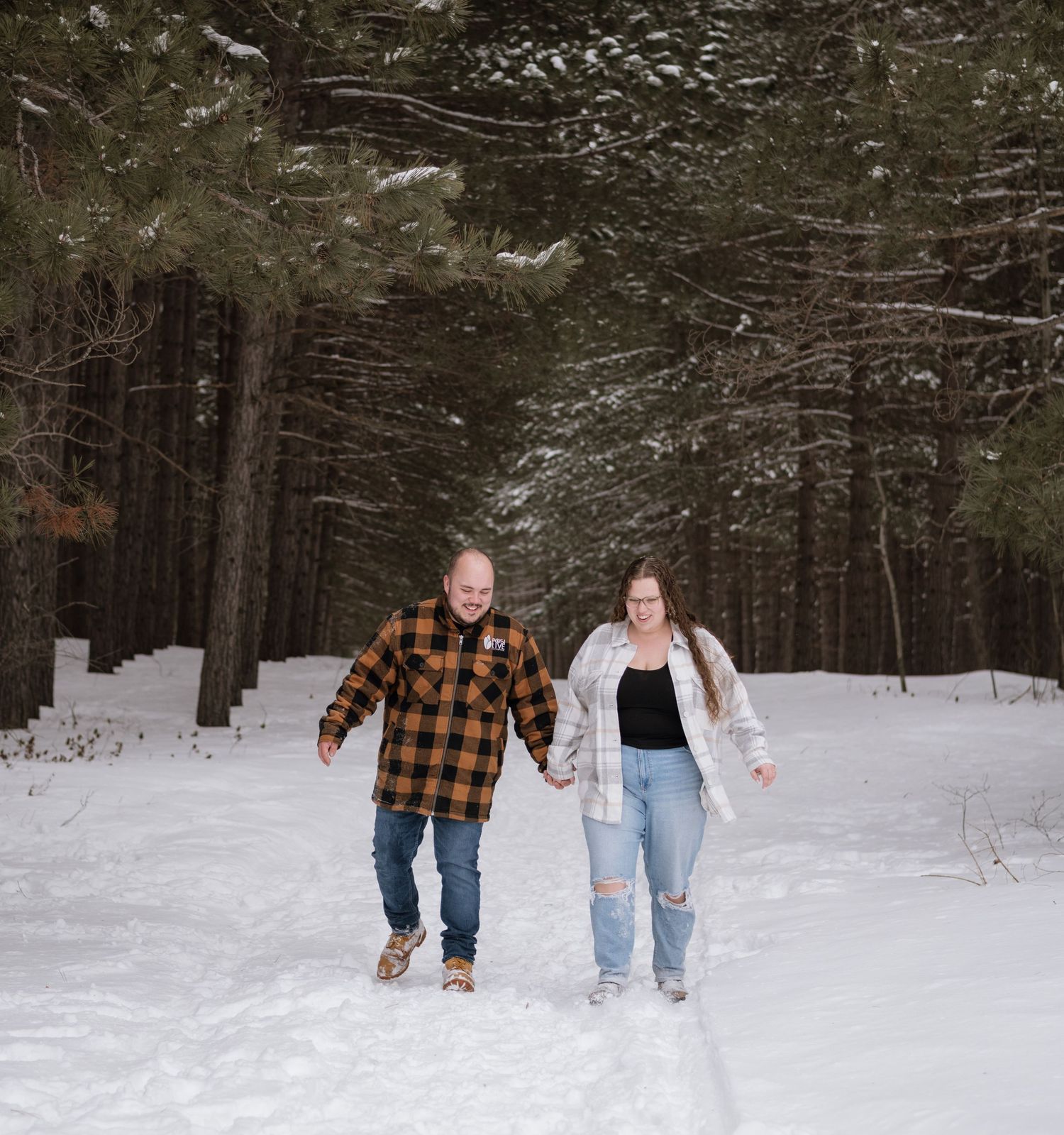 Couple holding hands walking through snowy forest path in winter, man in plaid shirt and woman in white cardigan.