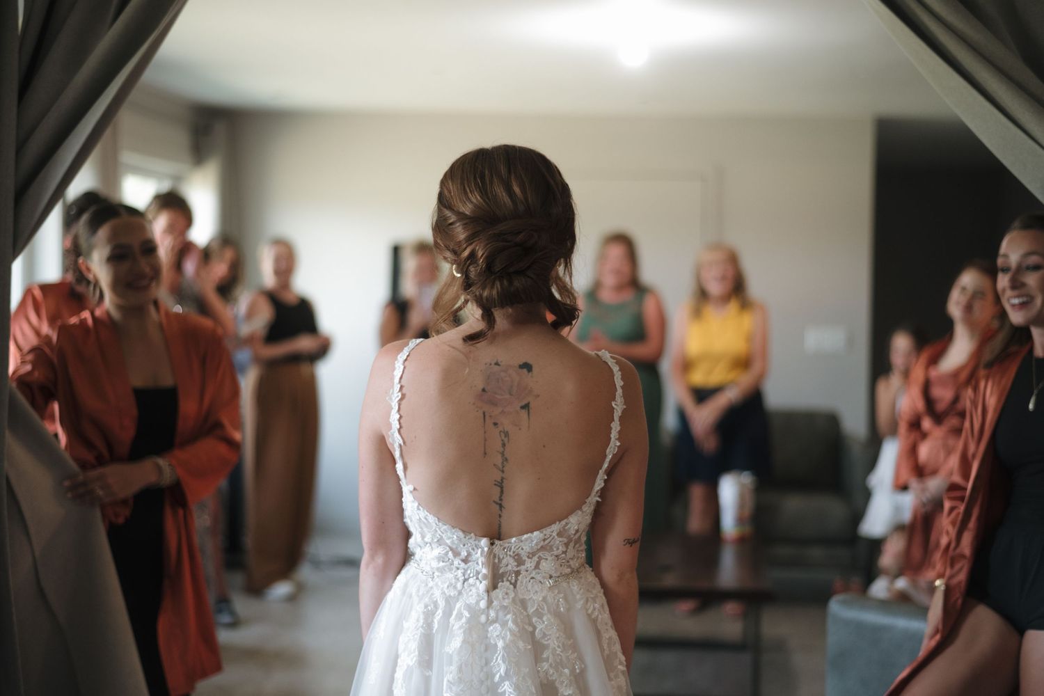 Bride in lace backless wedding dress with tattoo stands before bridesmaids in terracotta robes during bridal prep.