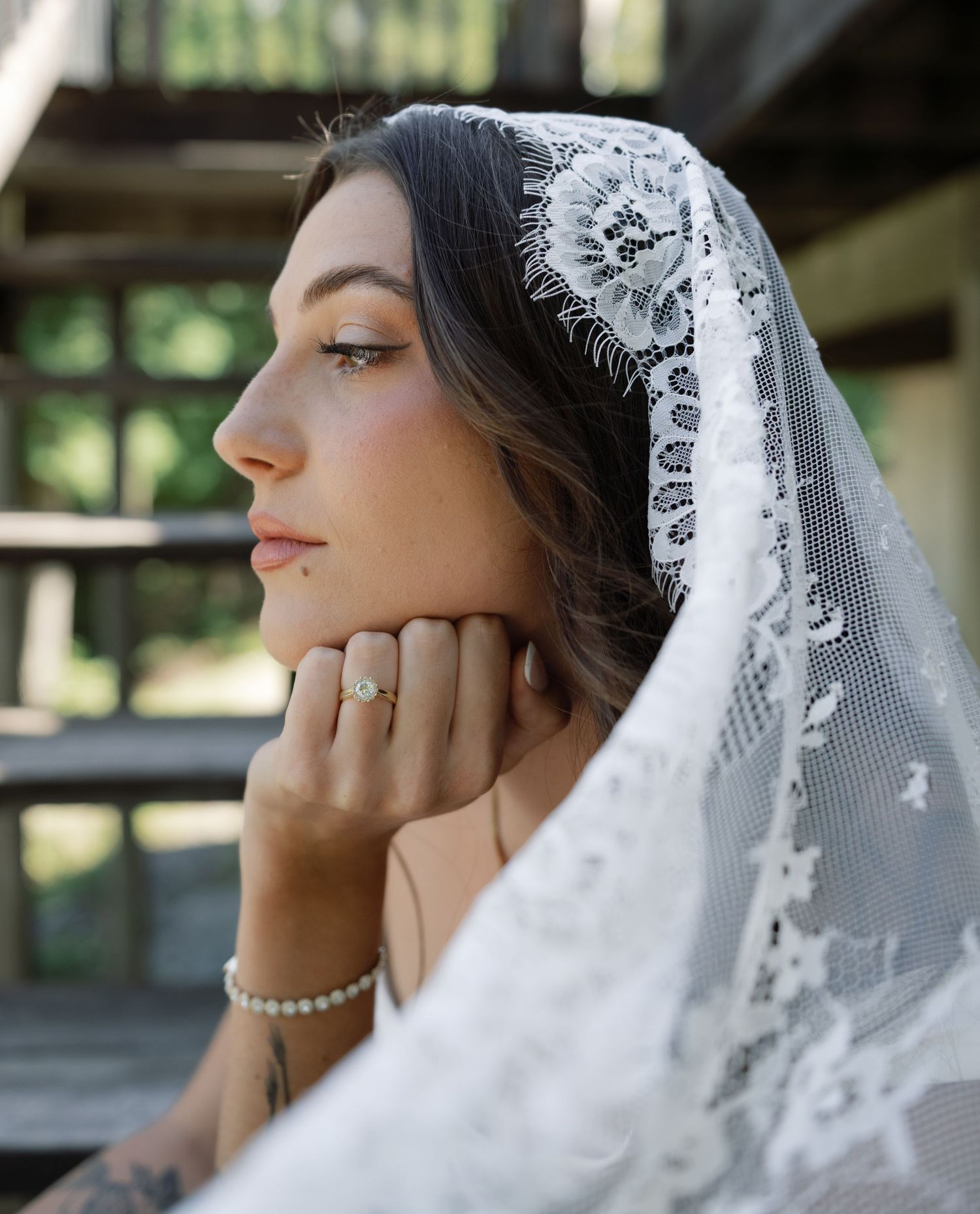Bride in white lace veil gazes thoughtfully, resting chin on hand, wearing pearl bracelet and ring outdoors.