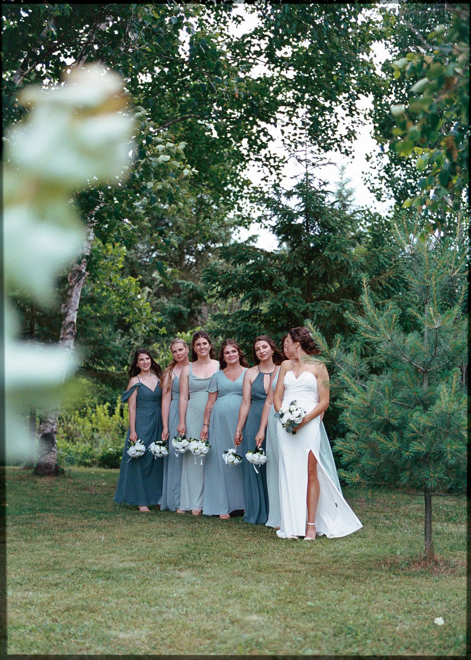 Bride in white gown stands with five bridesmaids in sage green dresses holding bouquets outdoors among lush green trees.