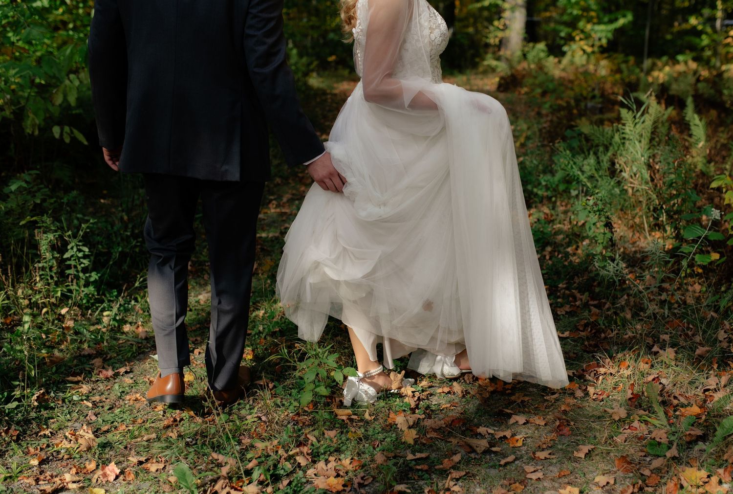 Bride and groom holding hands walking through a forest path, bride in white gown, groom in gray suit.