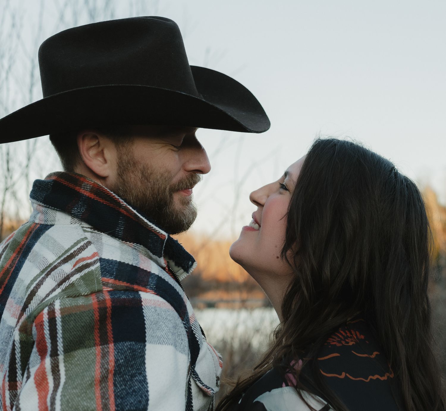 A couple in western attire share an intimate moment at sunset, with the person in a cowboy hat and plaid jacket.