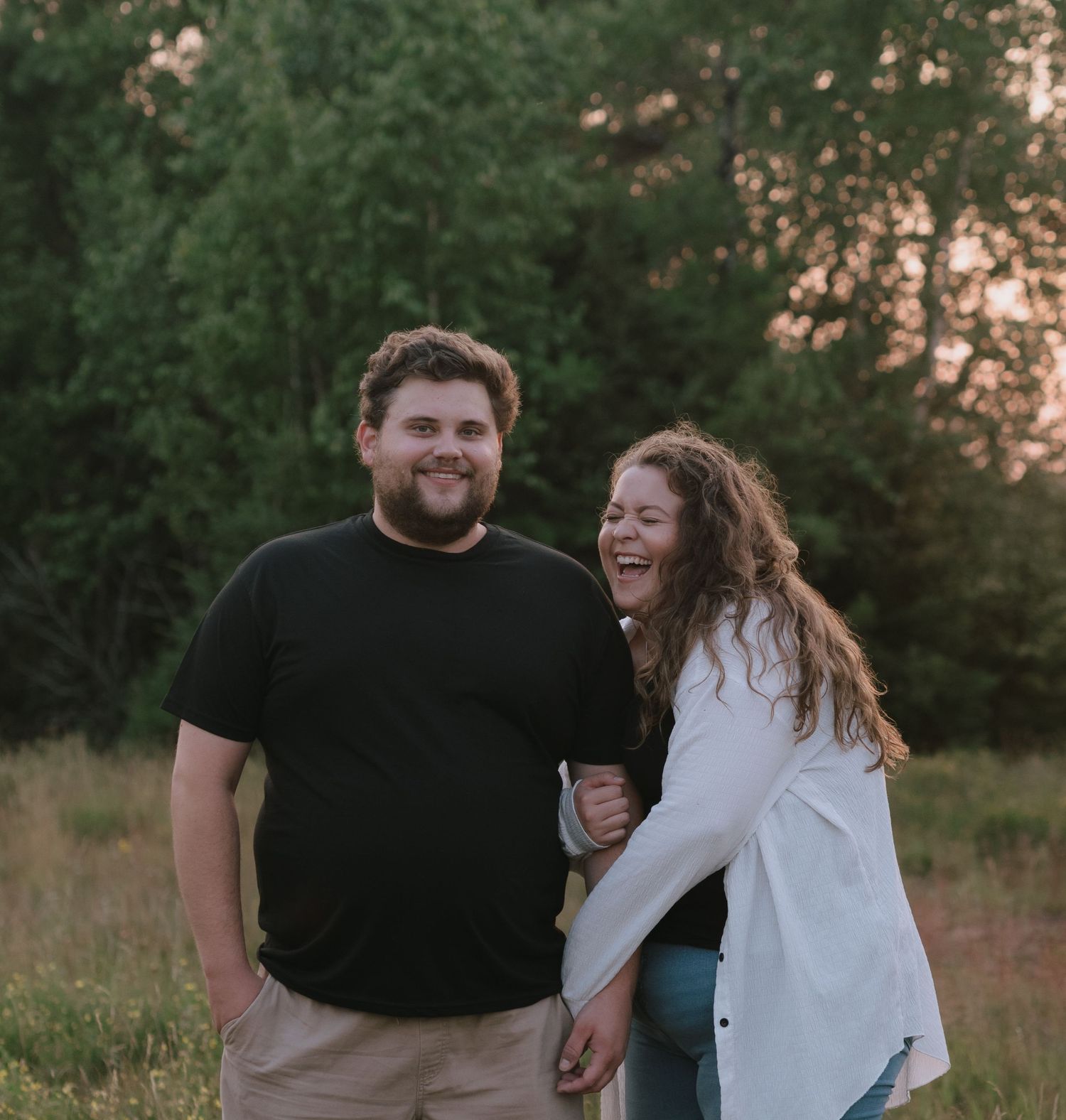 A happy couple laughs together in a field during golden hour.