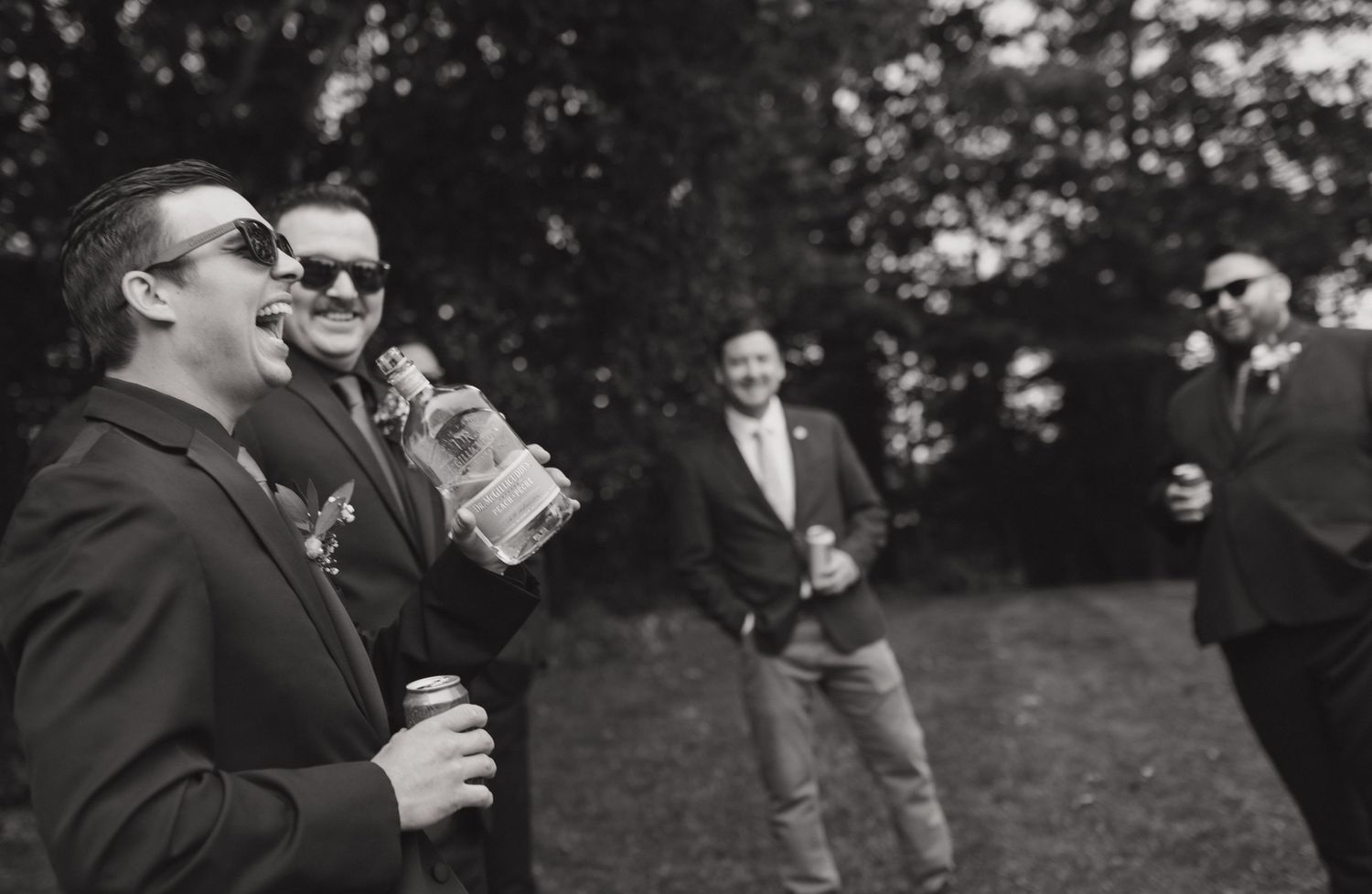 Black and white photo of groomsmen sharing drinks and laughs outdoors.