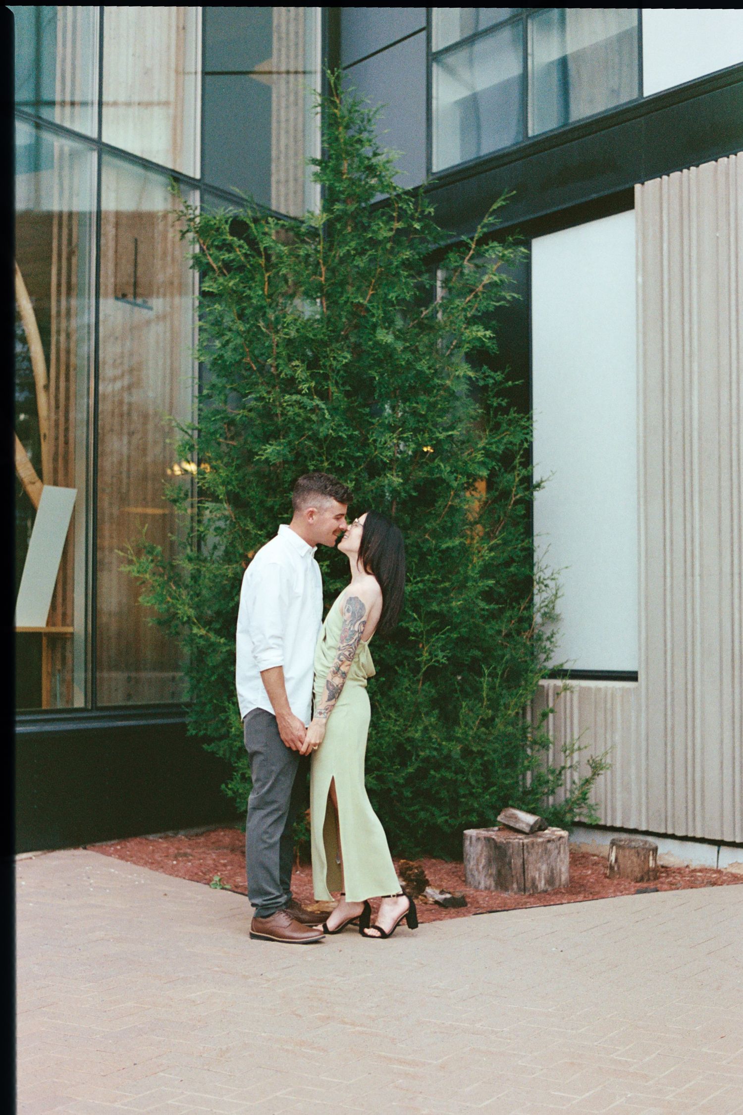 A couple in a romantic moment stands outside a modern building with glass windows and landscaping.