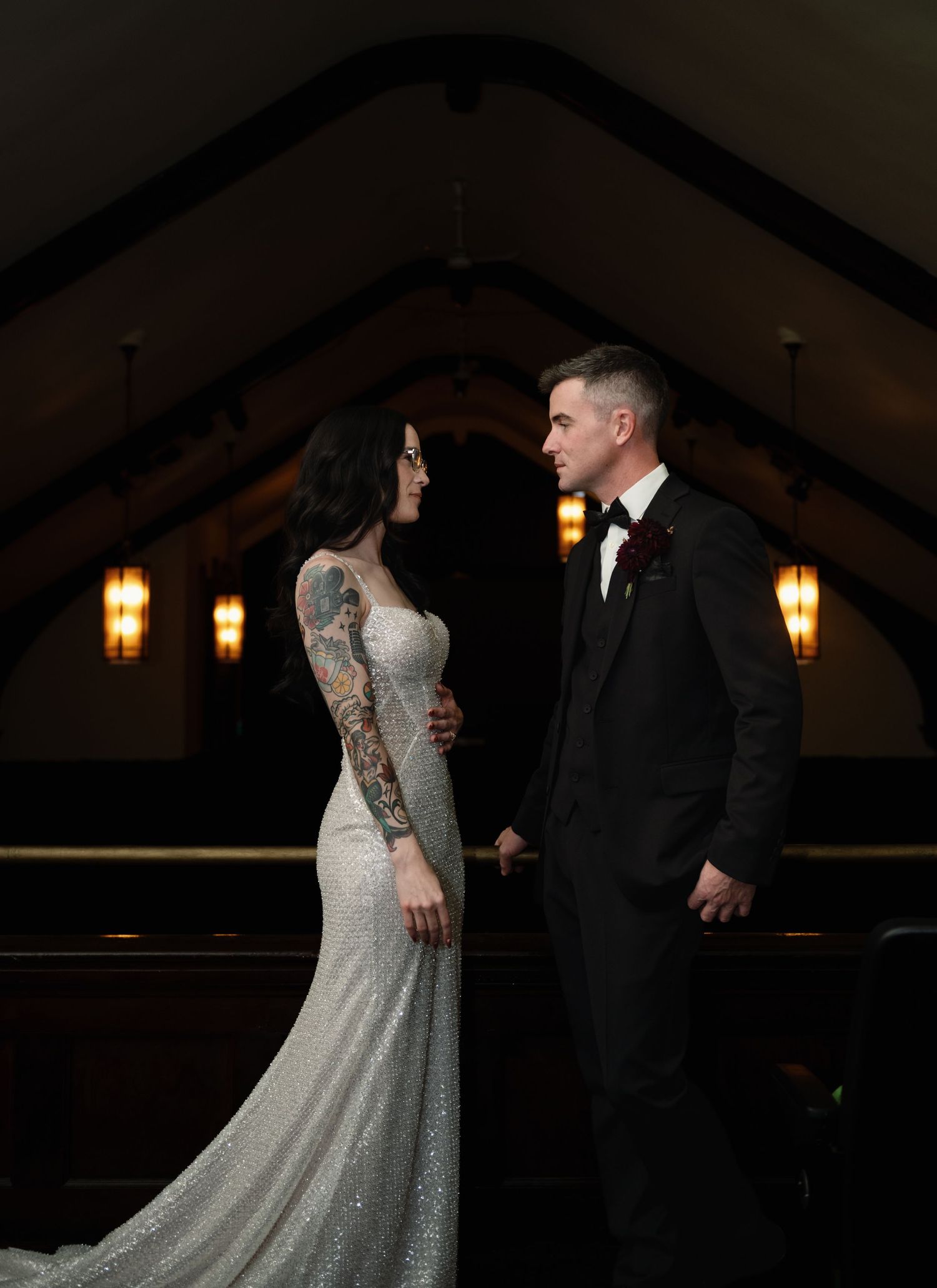 Bride in sparkly gown with sleeve tattoo and groom in black suit hold hands in a dimly lit chapel.