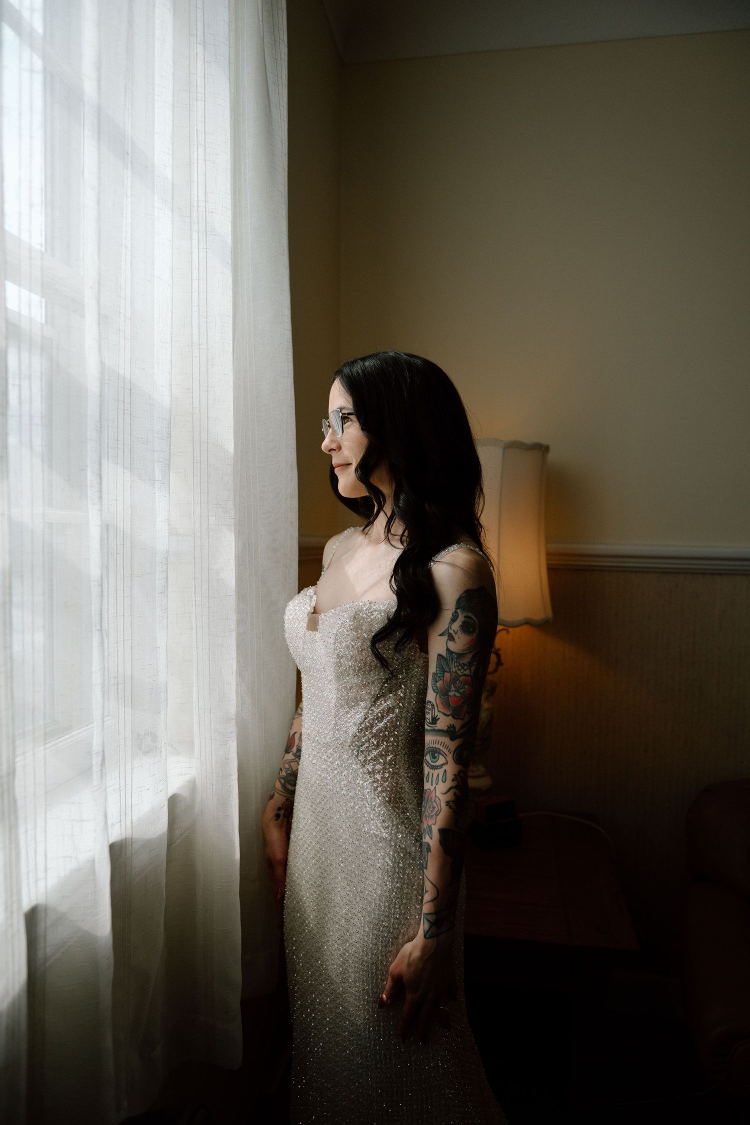 Tattooed bride in a fitted beaded gown standing by sheer curtains in soft natural light.