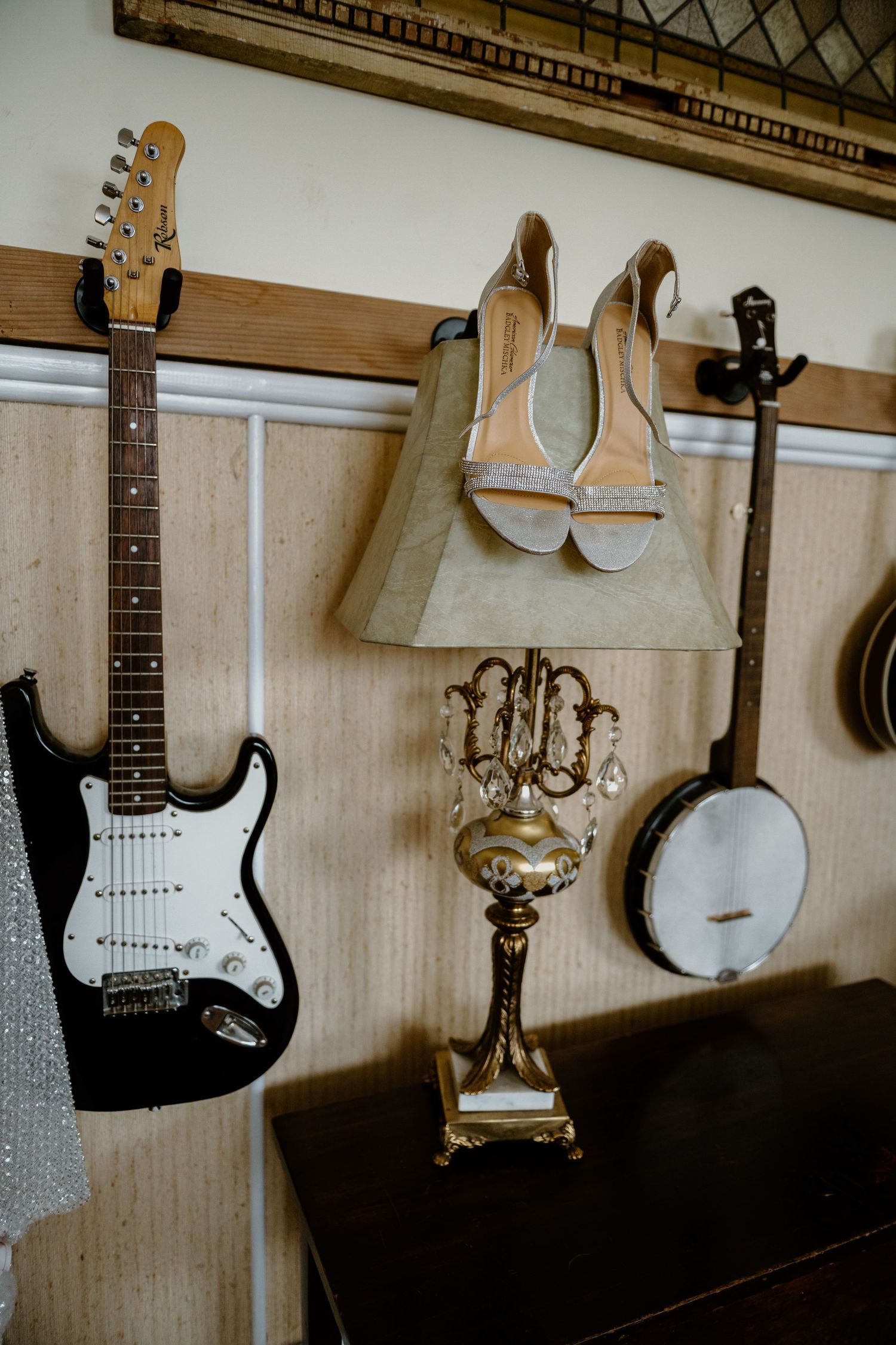 Vintage room decor featuring a black electric guitar, banjo, ornate gold lamp, and beige heels displayed on a wooden wall.
