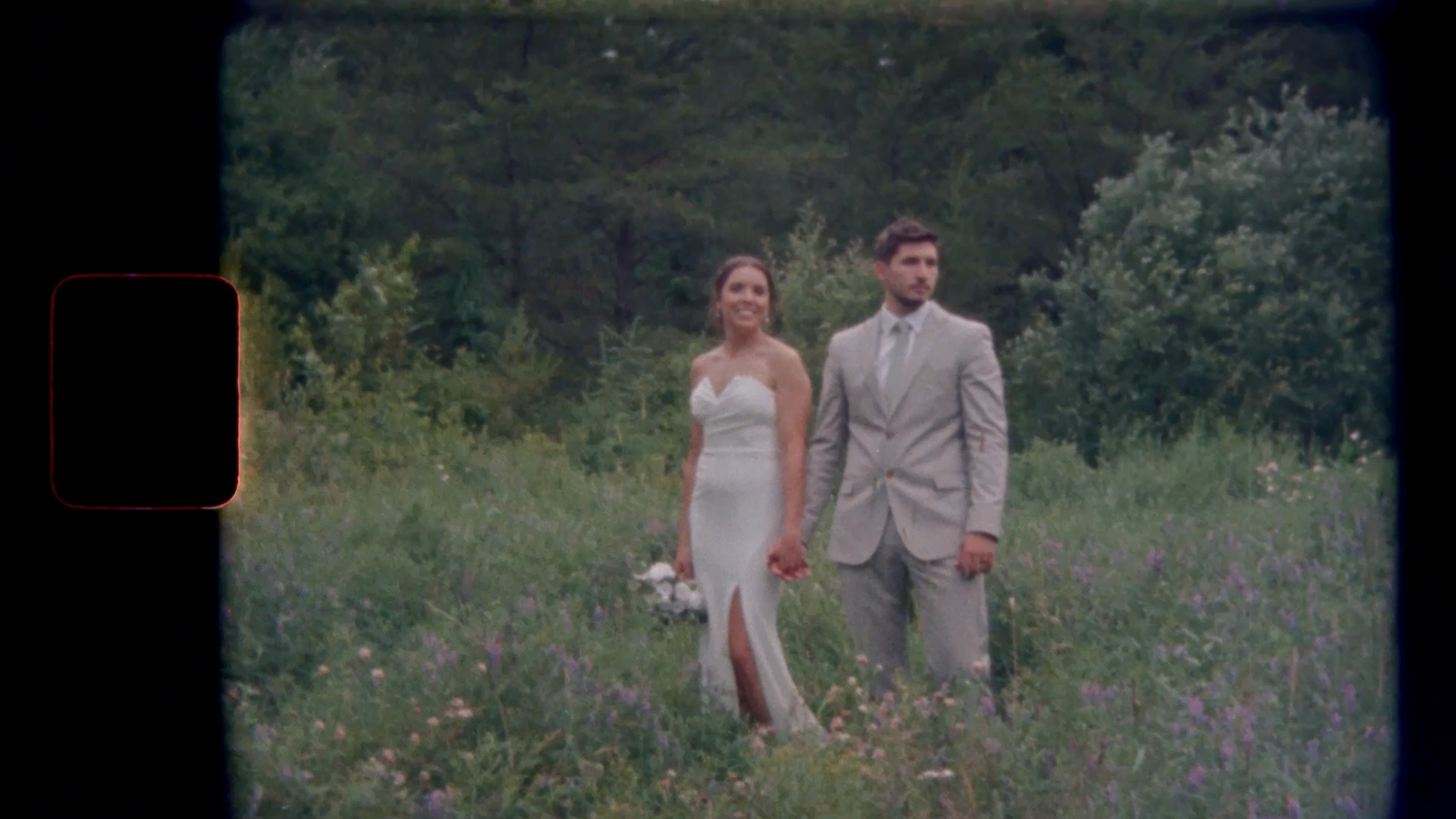 A bride and groom stand together in a wildflower meadow, she in a white gown, he in a tan suit, with lush greenery behind.