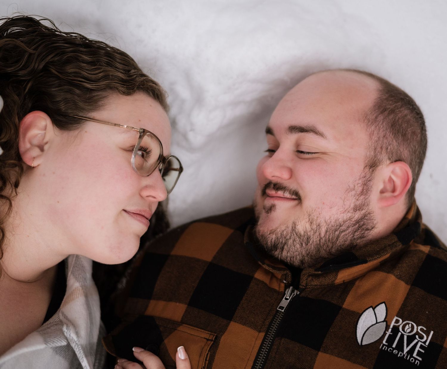Couple lying in snow, smiling at each other, woman with glasses, man in brown plaid jacket with logo.