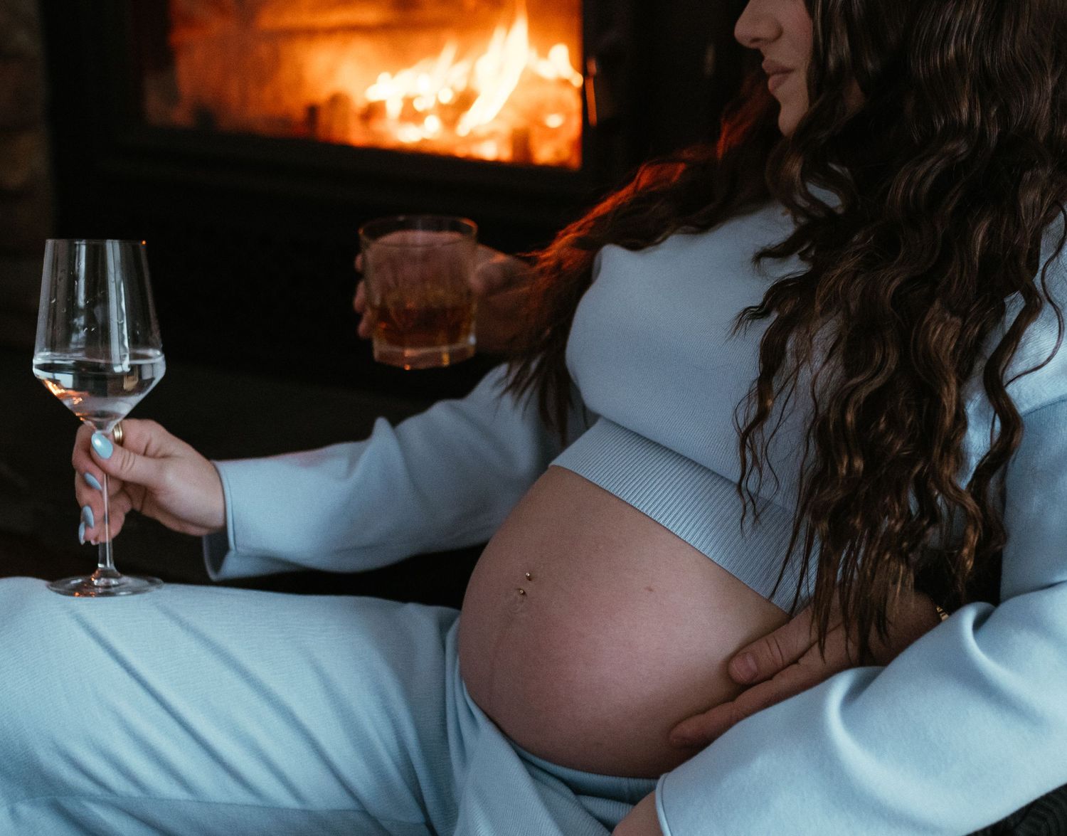 Pregnant woman in light blue outfit holding wine glass by fireplace during maternity session.