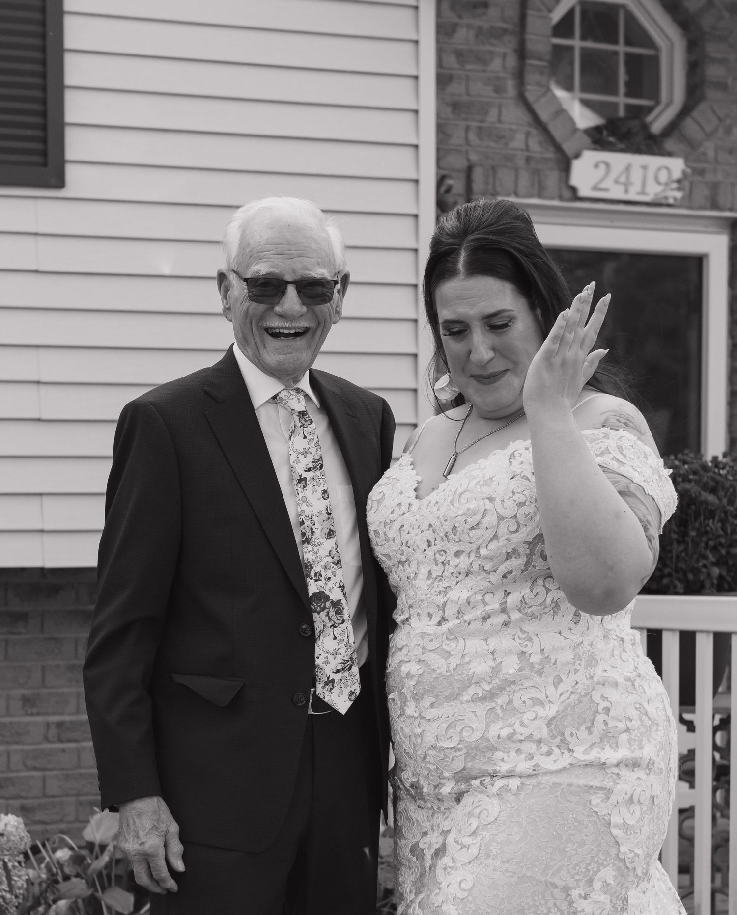Black and white photo of a bride in lace gown showing her ring beside an elderly man in suit at a house.