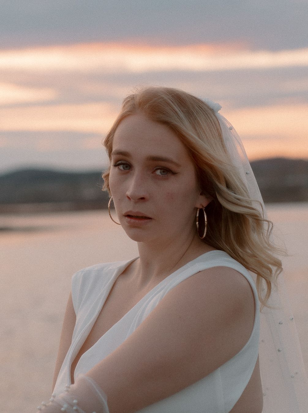 A woman in white poses by the water during sunset with soft pink sky in the background.