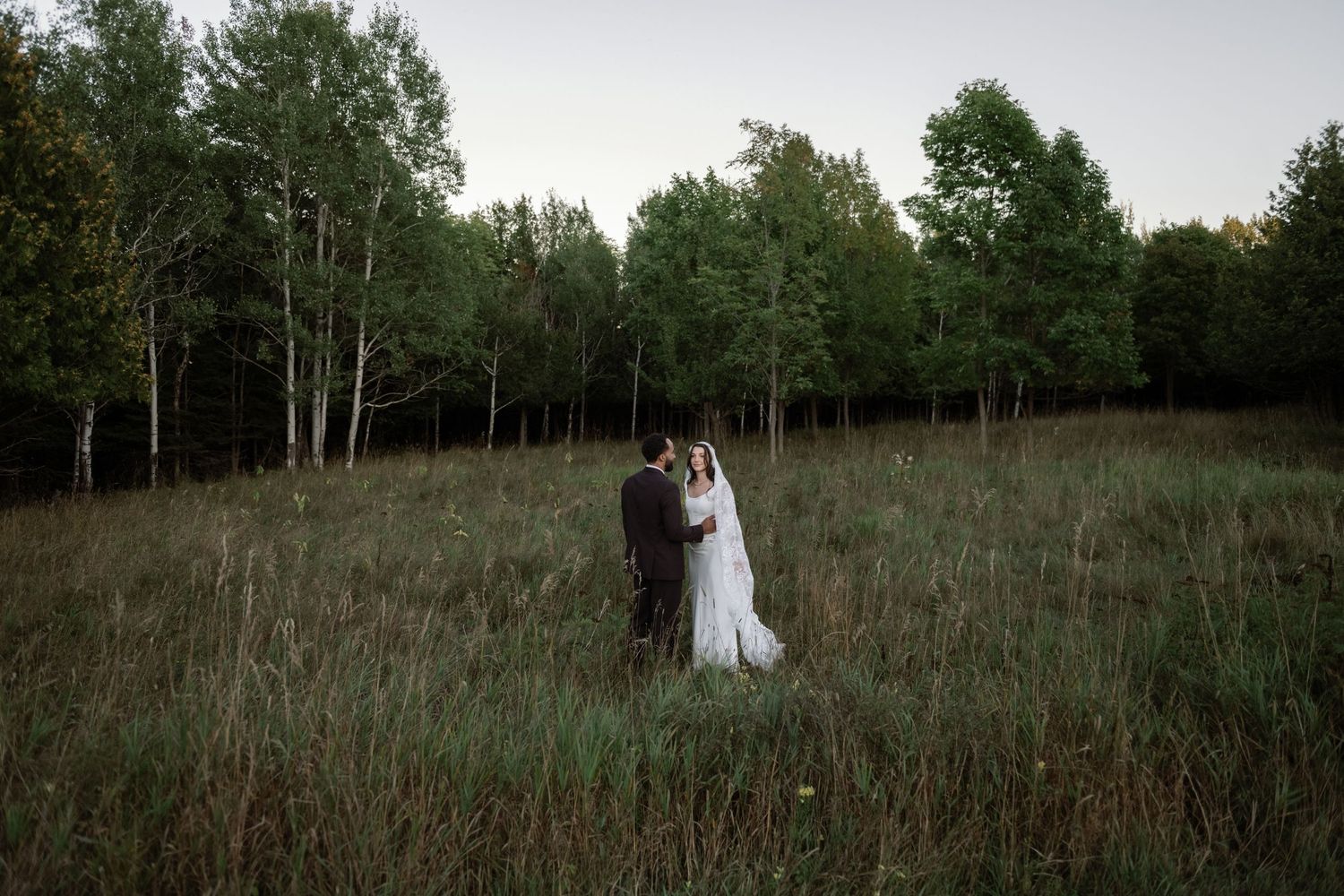 Bride and groom stand together in a lush meadow surrounded by tall trees at twilight.