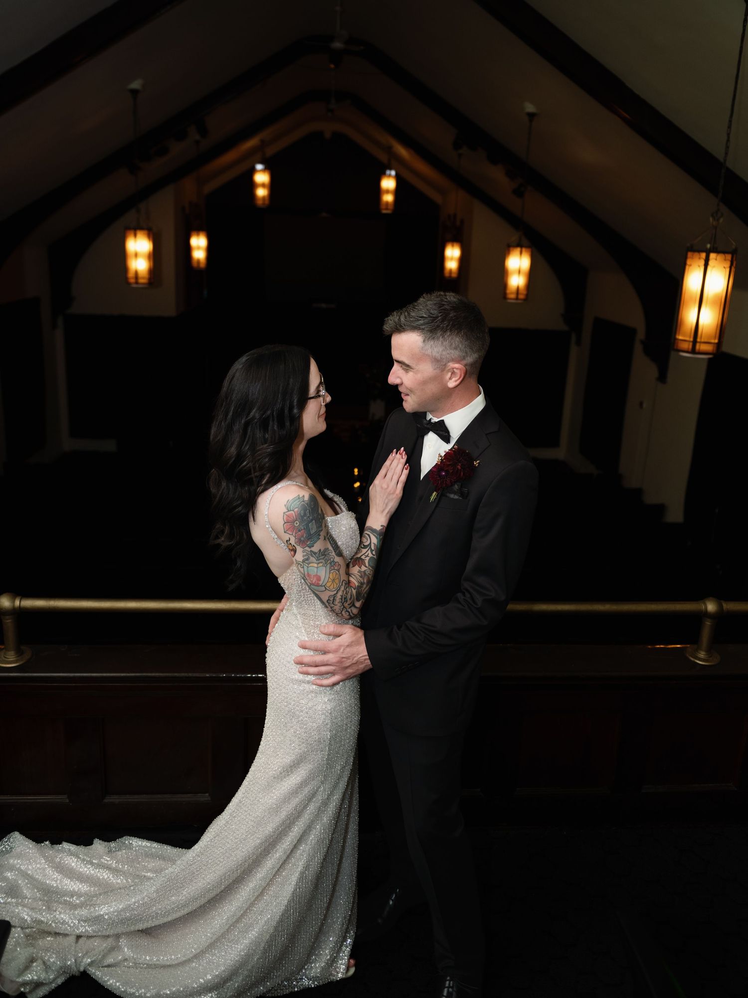 Bride and groom share a romantic moment in a dimly lit chapel, bride wearing a lace gown with tattoos visible.