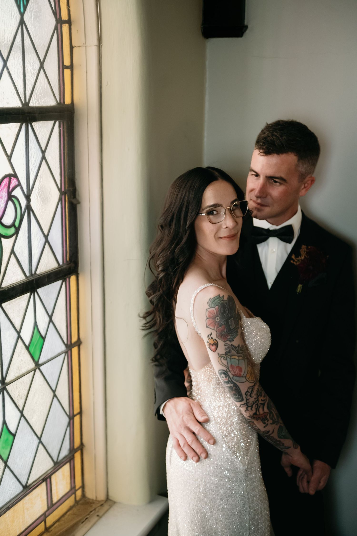 Bride with tattoos in sparkly gown and groom in black tuxedo pose by stained glass window in romantic wedding portrait.