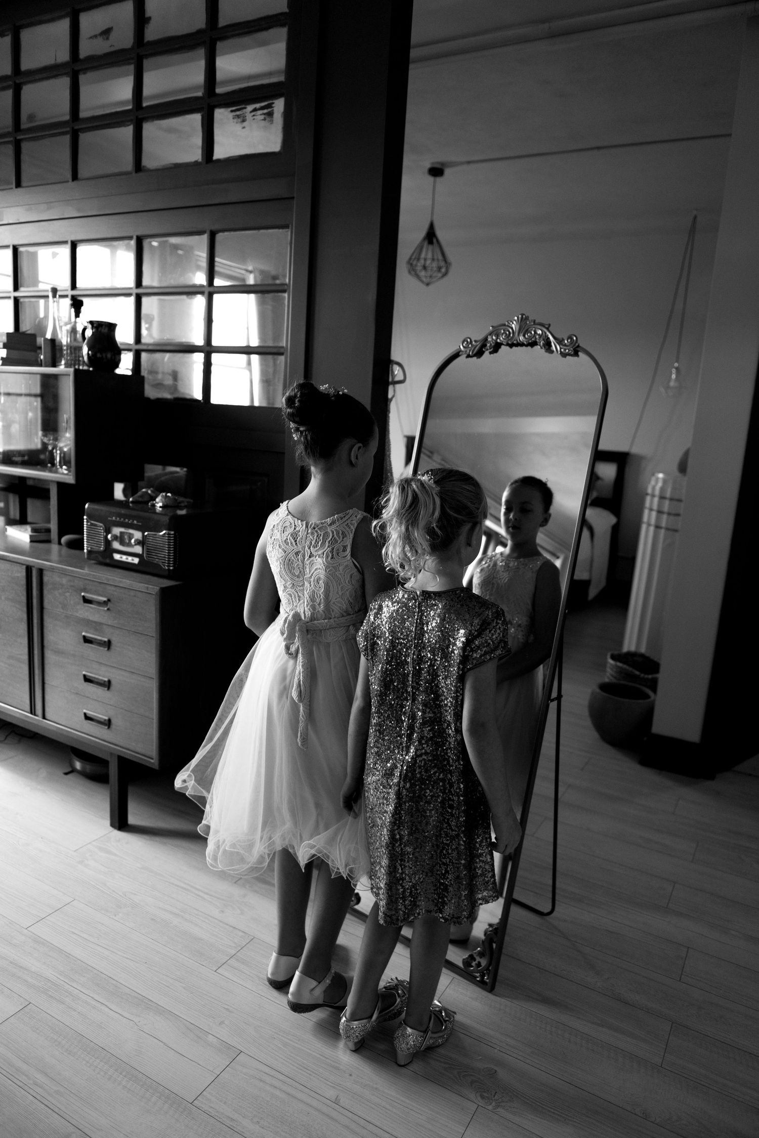 Two girls in formal dresses admire their reflections in an ornate mirror, captured in black and white.