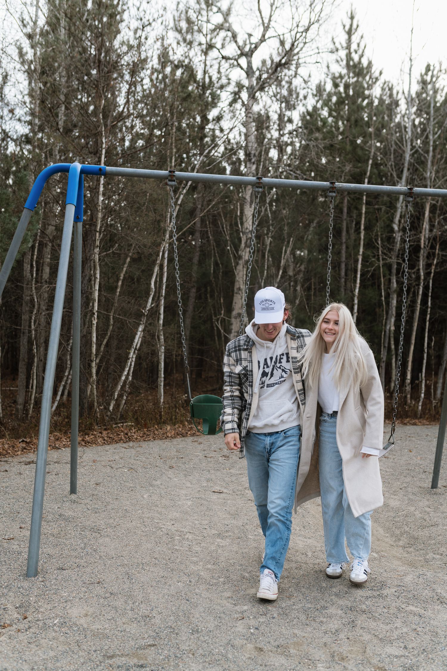 Couple walking and laughing together near a swing set in a wooded park on a cloudy day.