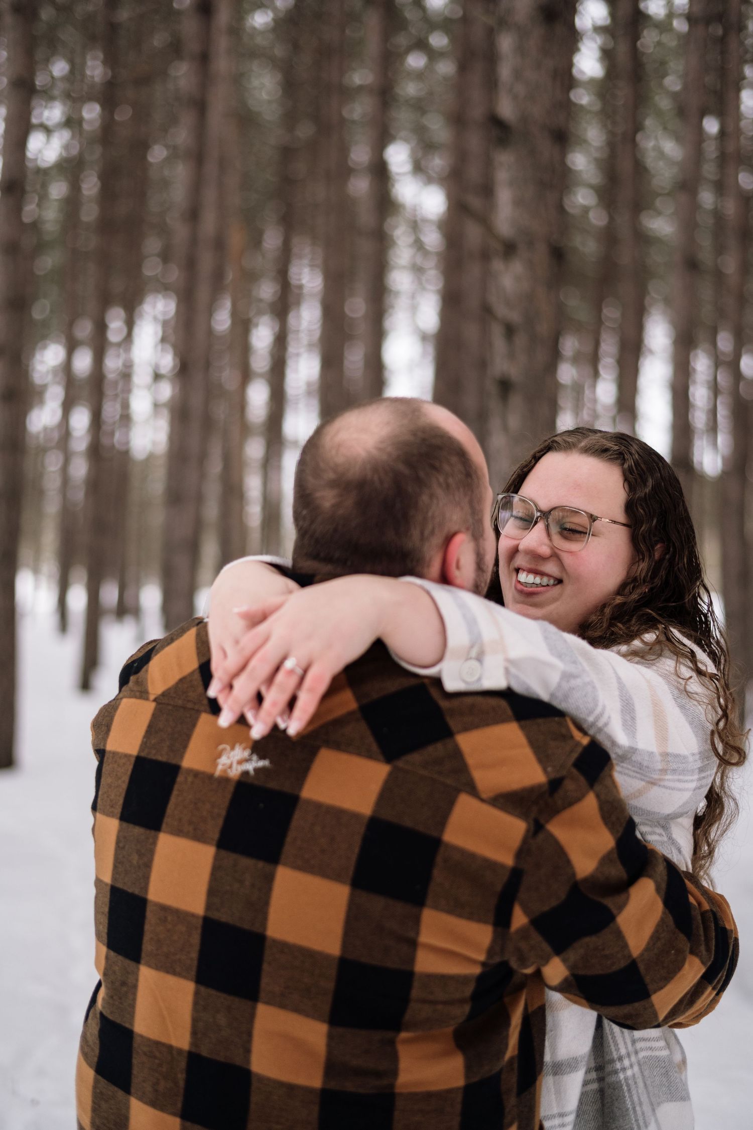 Couple embracing and laughing in snowy pine forest, woman wearing glasses and white top, man in brown plaid flannel jacket.