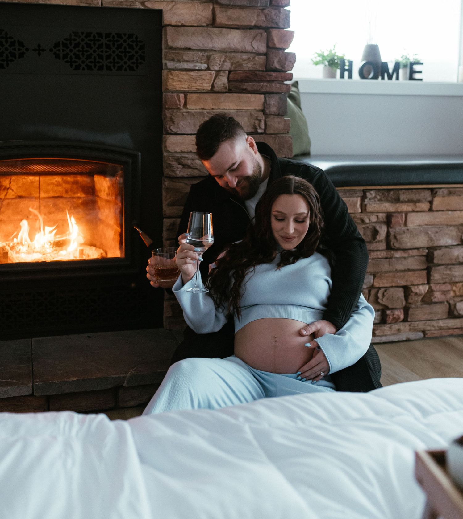 Expecting couple sitting by a warm stone fireplace, man holding pregnant womans belly.