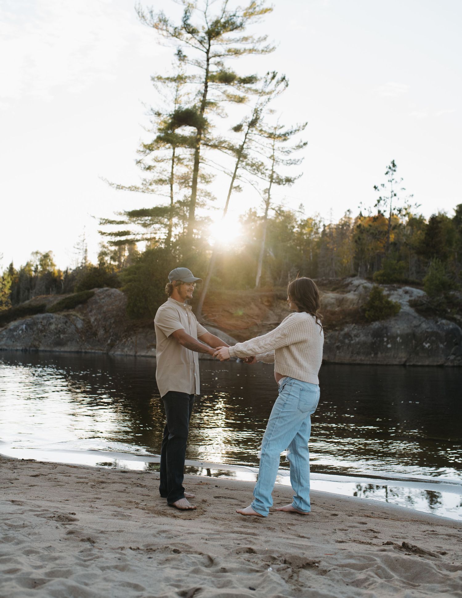 A couple dances together on a sandy beach at sunset near a lake.
