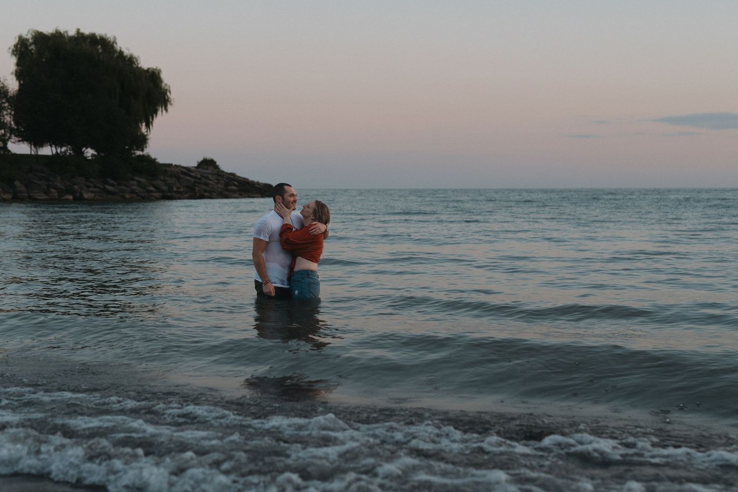 A couple stands together in shallow ocean water during a peaceful sunset.