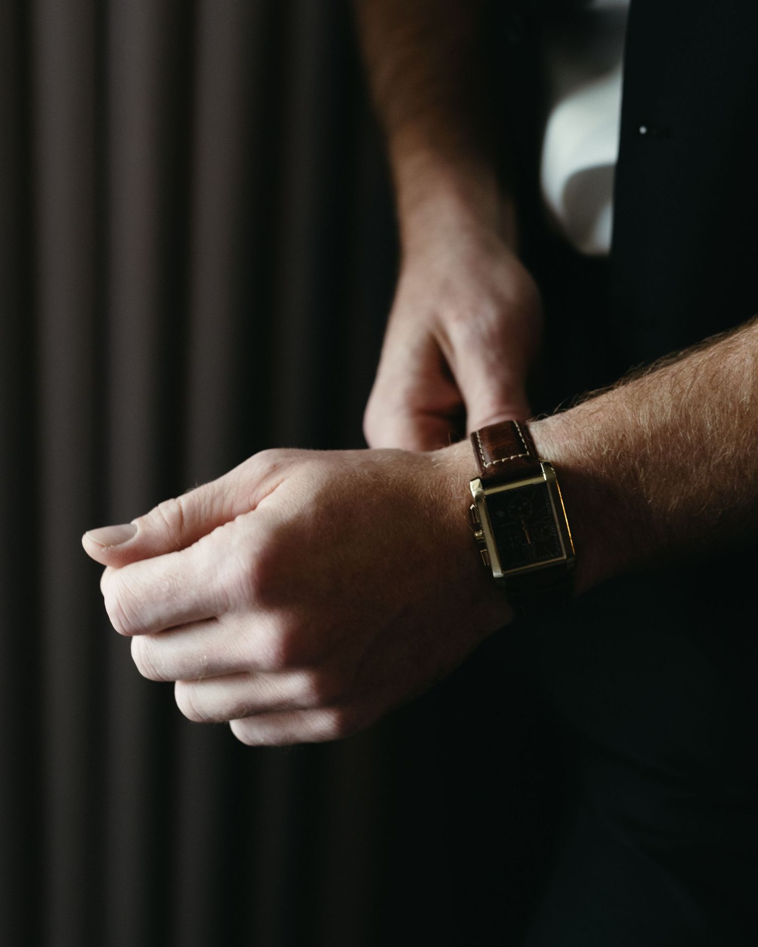 Close-up shot of hands adjusting a leather watch with gold accents against a dark background.