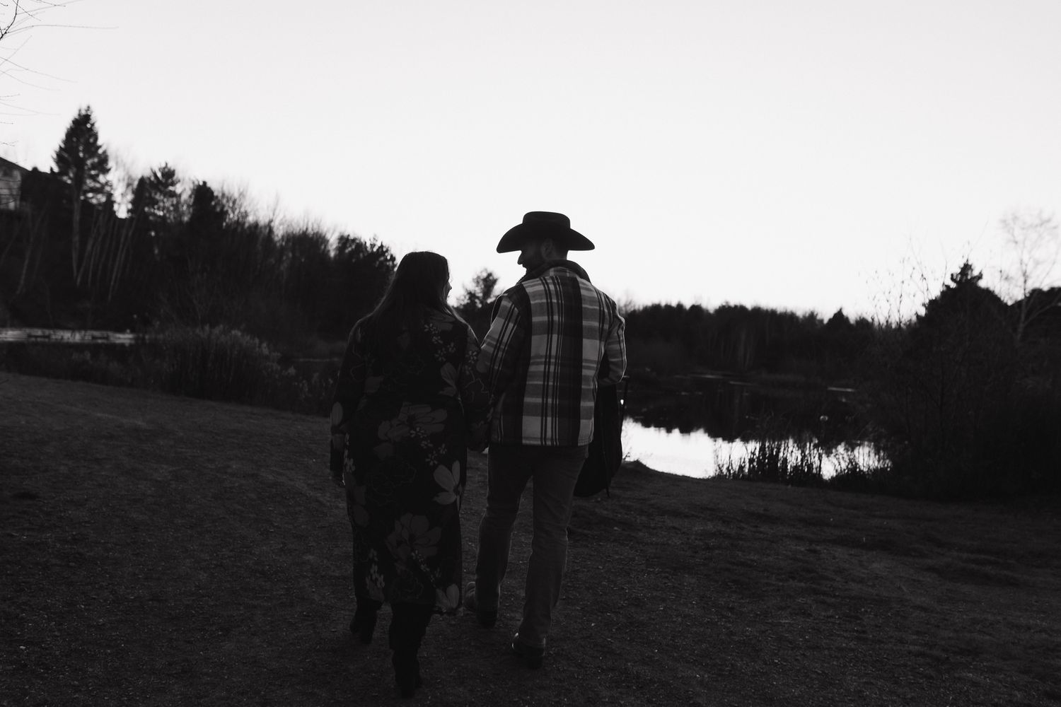 Black and white silhouette of two people walking near a lake at dusk with trees in the background.