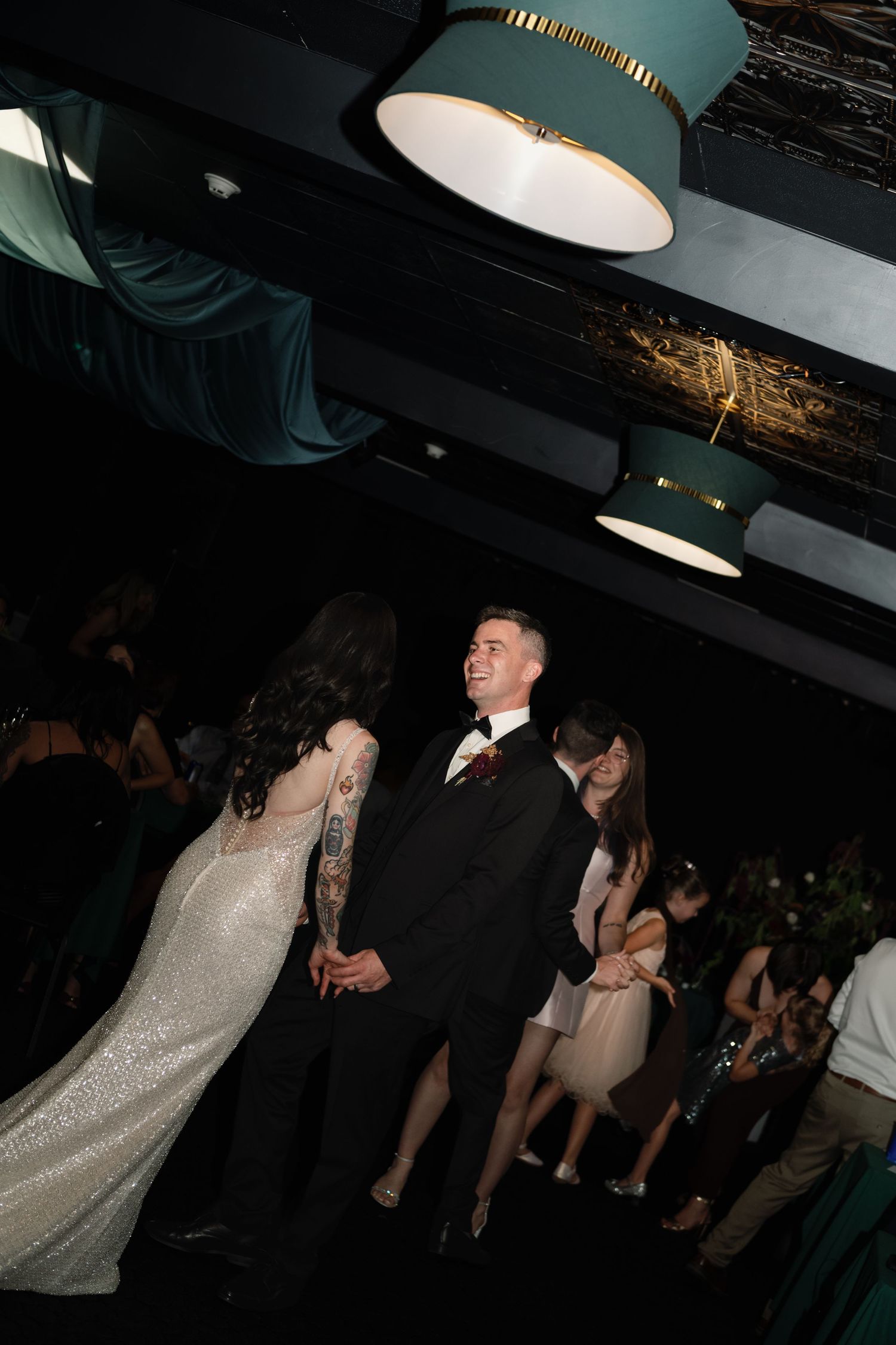 A smiling groom in a black tuxedo shares a dance with his bride in a sequined gown at an elegant wedding reception.