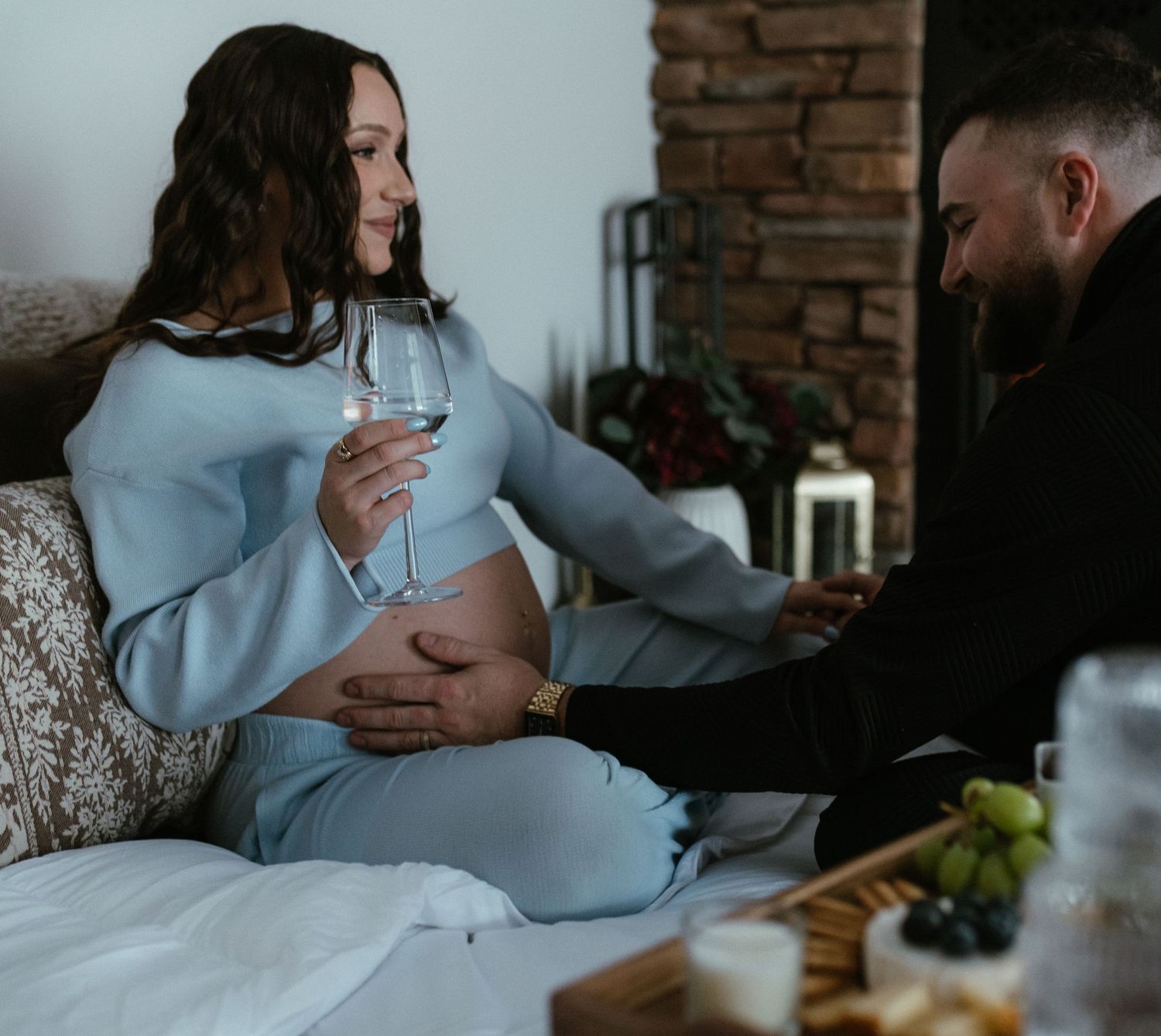 Pregnant woman in blue outfit holds wine glass while partner tenderly touches her baby bump during intimate maternity session.