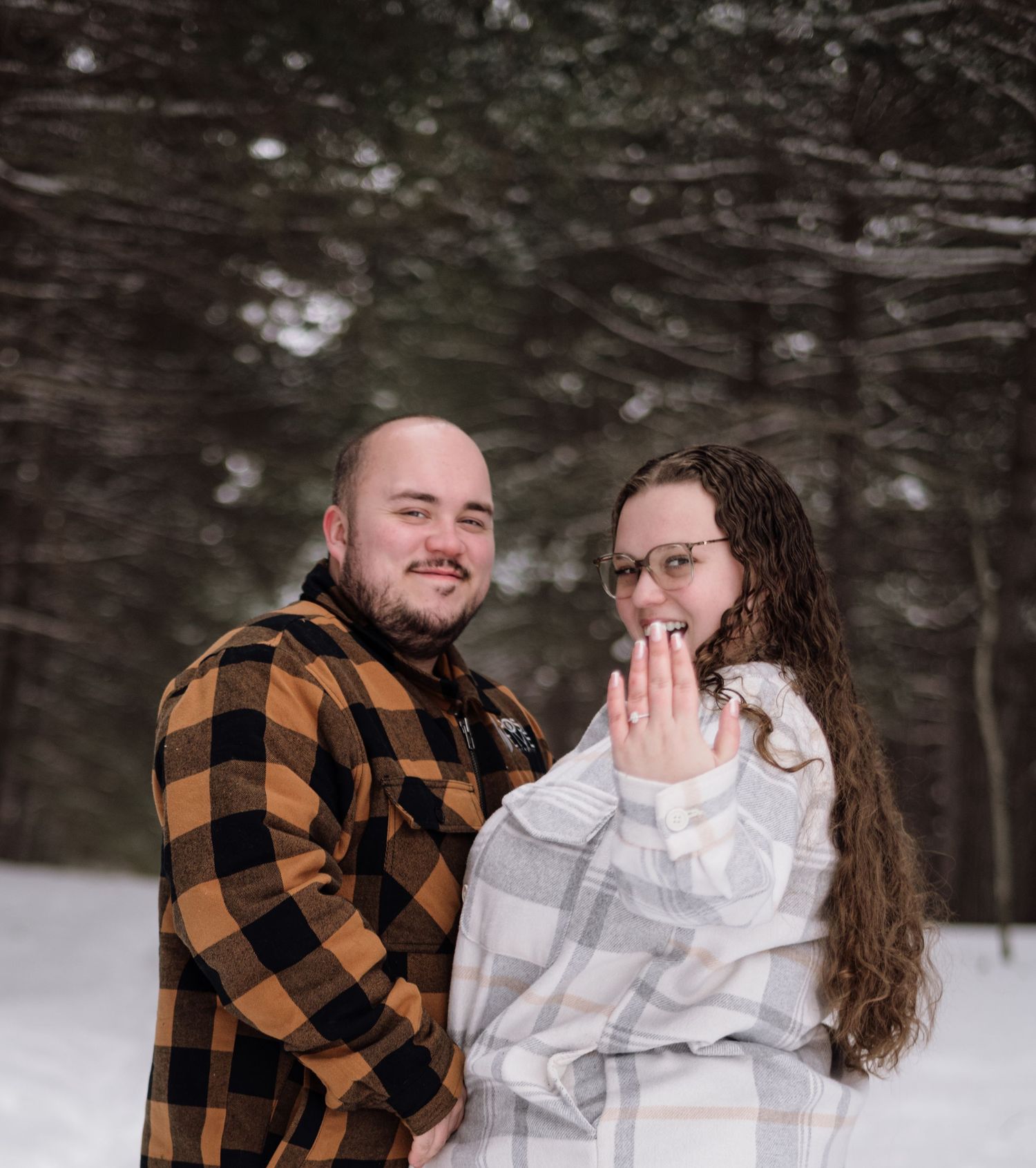 Couple posing in snowy forest, woman showing engagement ring, man in plaid flannel shirt.