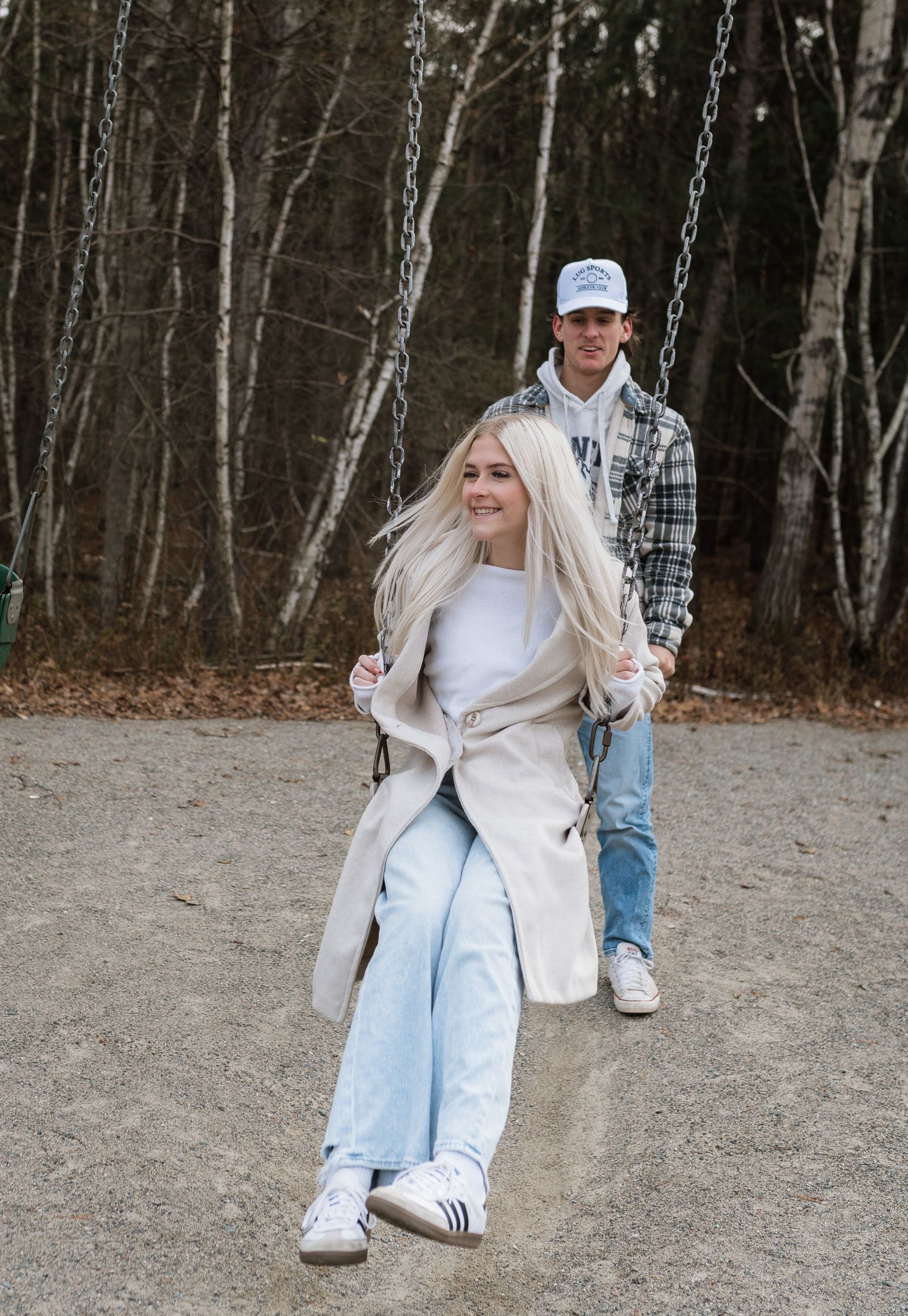Young woman with blonde hair sitting on a swing while a man in a plaid shirt pushes her at an outdoor playground.