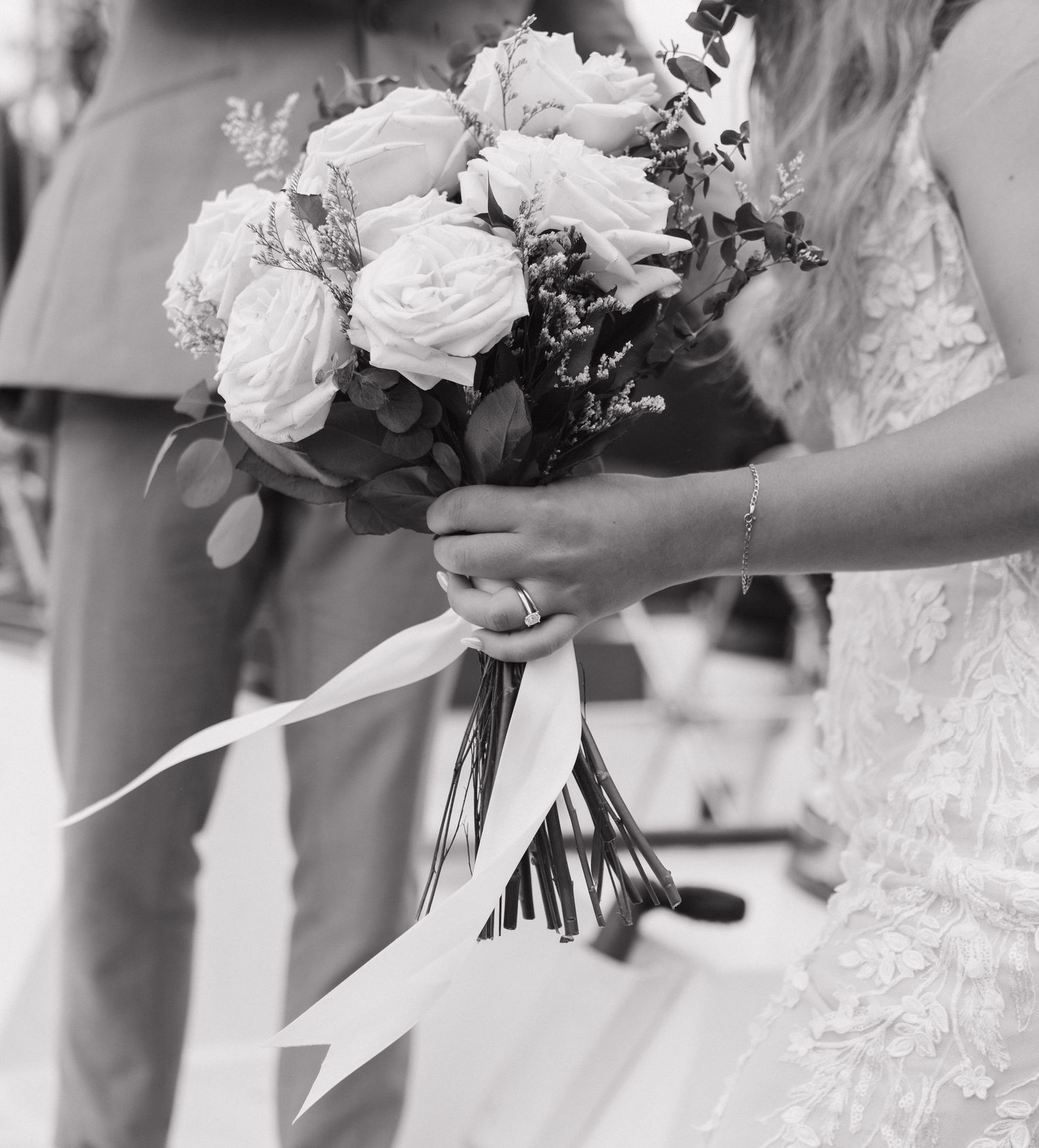 Black and white photo of a bride holding a lush rose bouquet with ribbon streamers at a wedding ceremony.