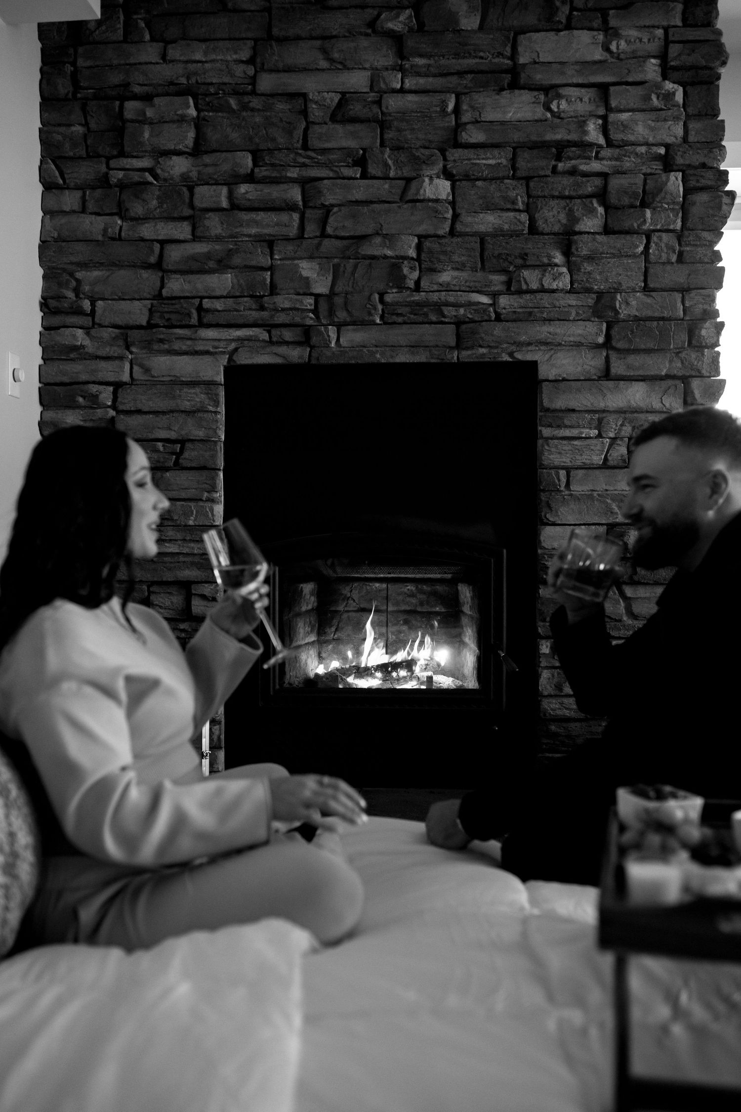 Black and white photo of a couple drinking wine by a stone fireplace while sitting on a bed.