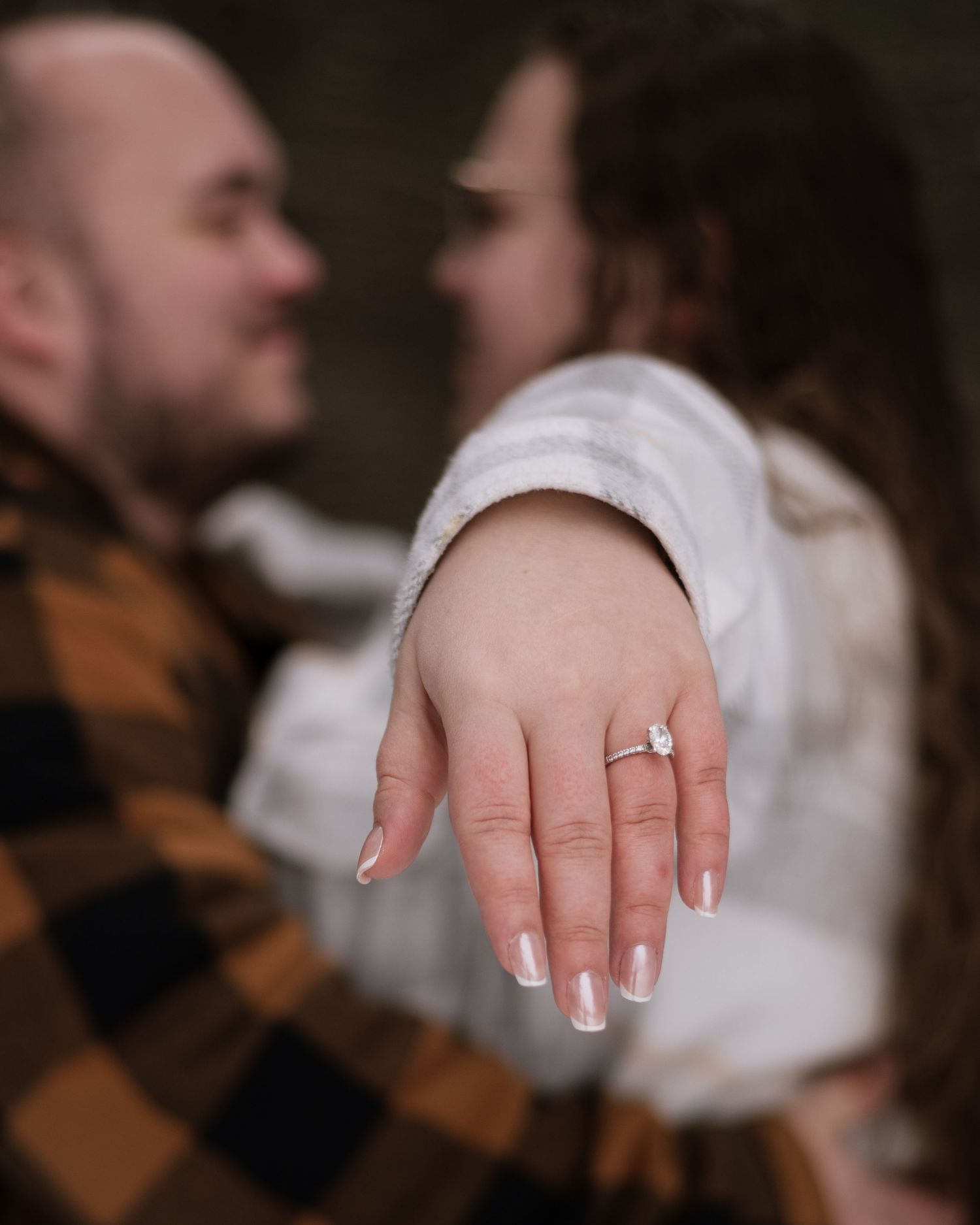 Couple sharing an engagement moment, woman displaying diamond ring on finger, man in plaid shirt smiling in soft bokeh background.