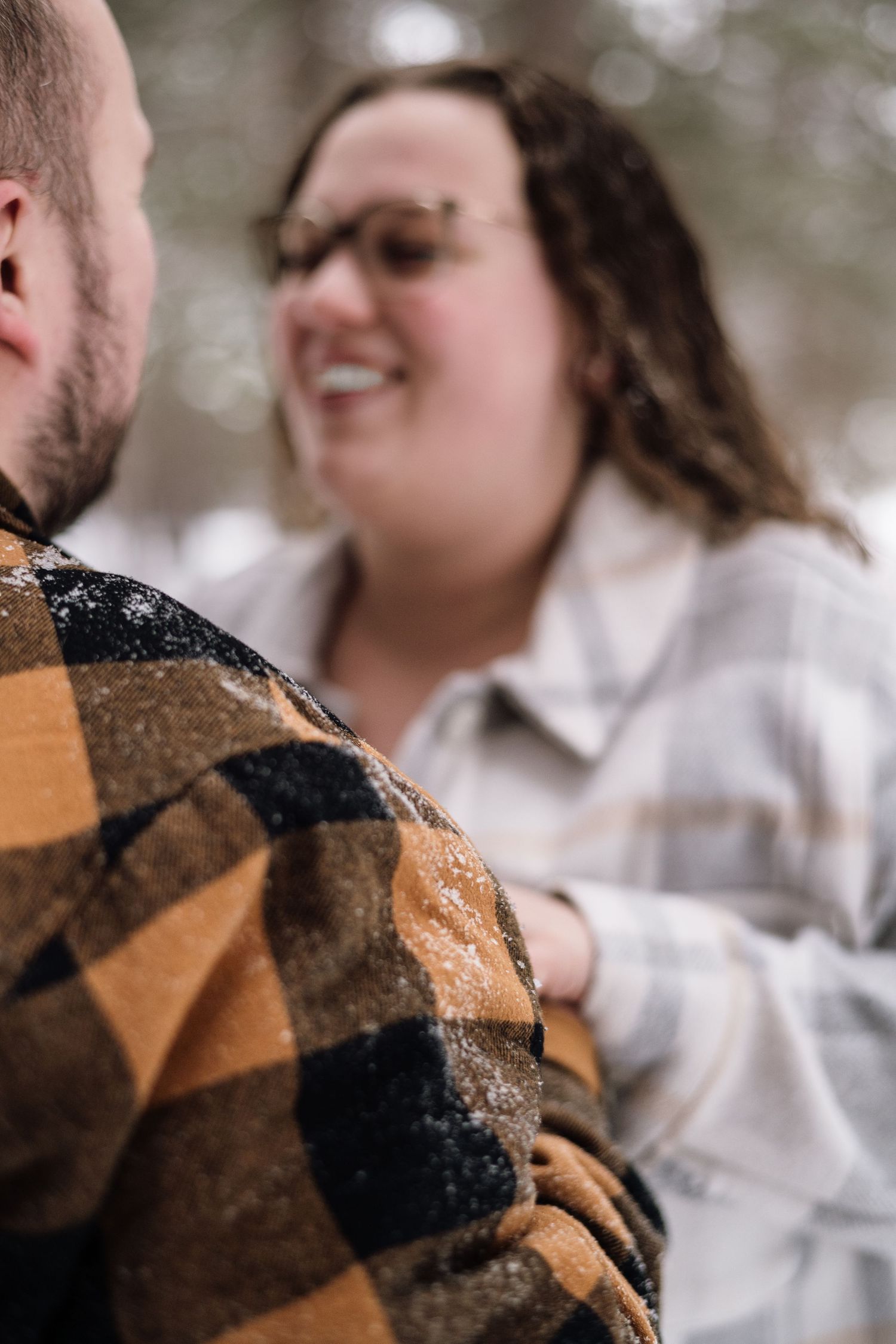Smiling woman in white plaid jacket laughs while holding man in snow-dusted orange buffalo check flannel in winter forest.
