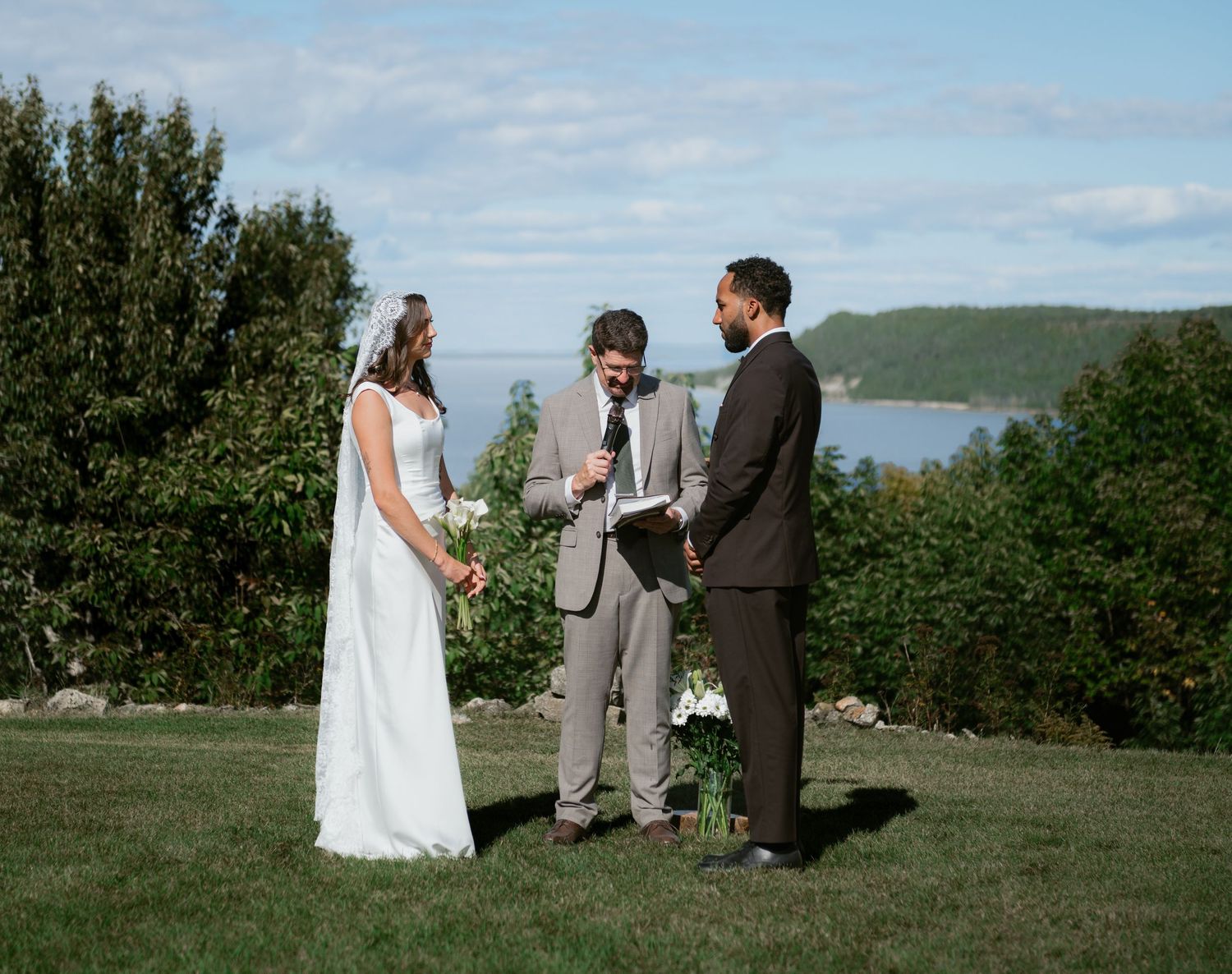 Outdoor wedding ceremony on green lawn with bride, groom, and officiant, scenic lake and hills in background.