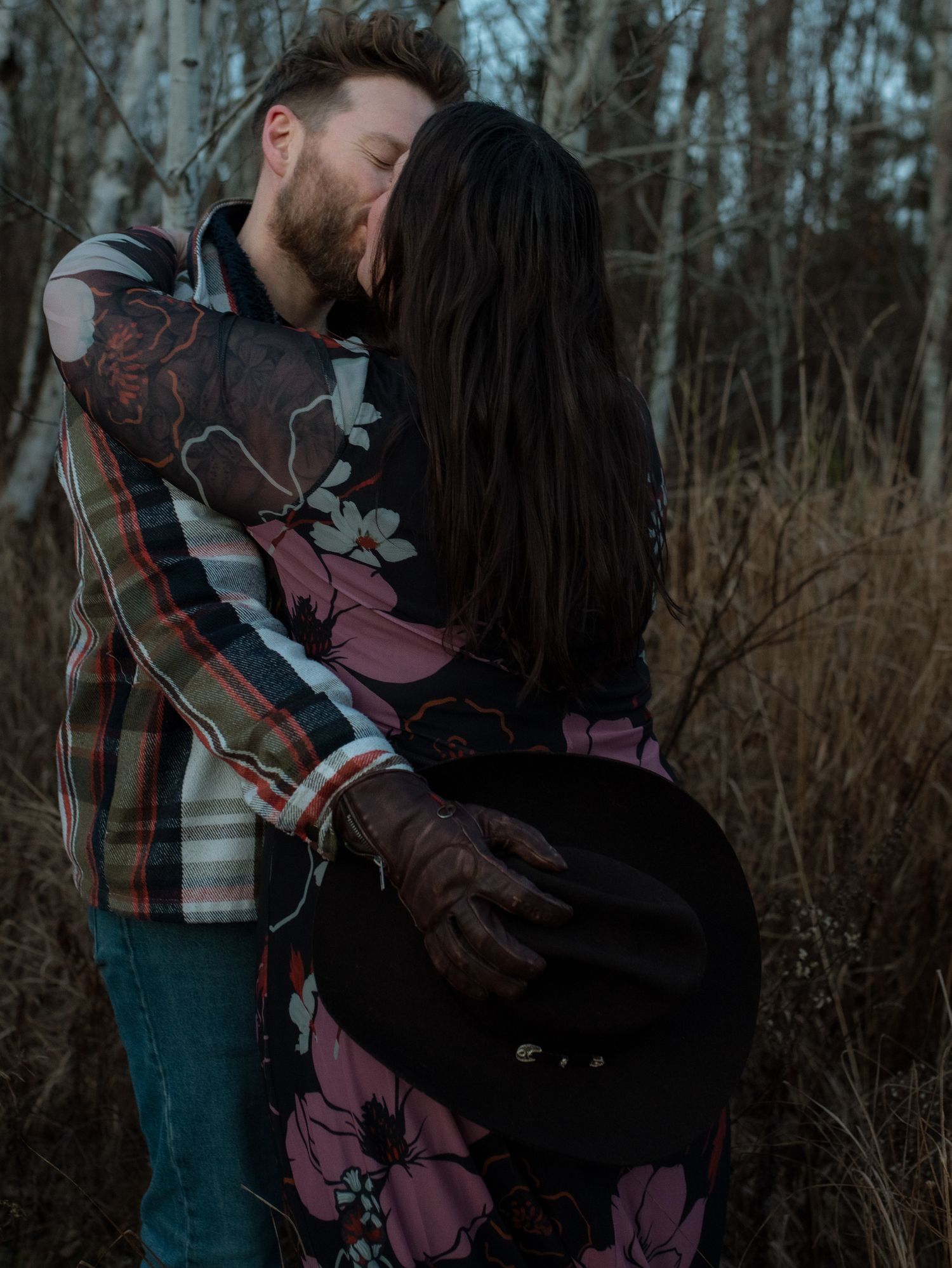A couple shares an intimate embrace in a moody forest setting with dramatic lighting and muted autumn tones.