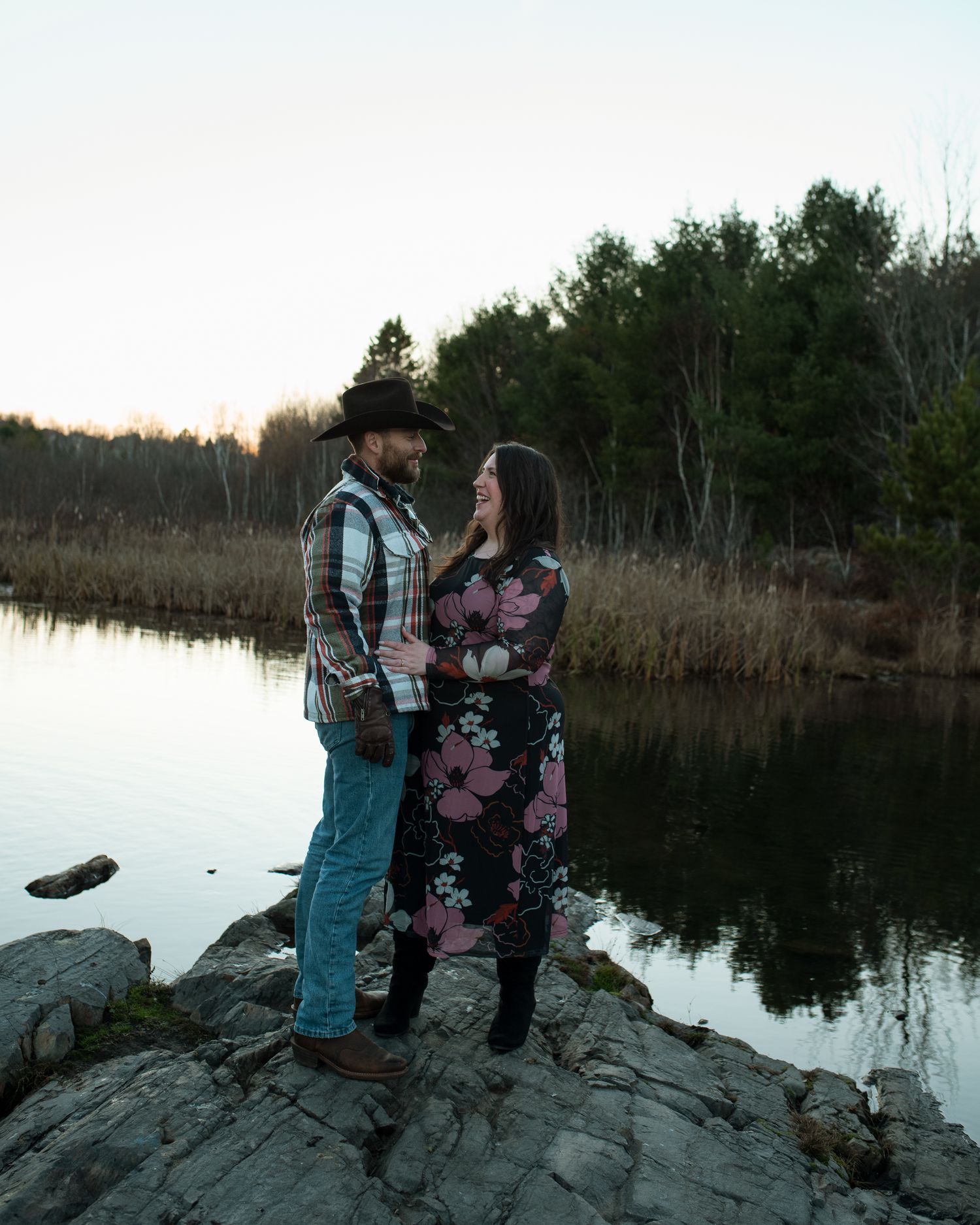 A couple stands together on a rocky outcrop by a serene lake at dusk, surrounded by pine trees and autumn foliage.