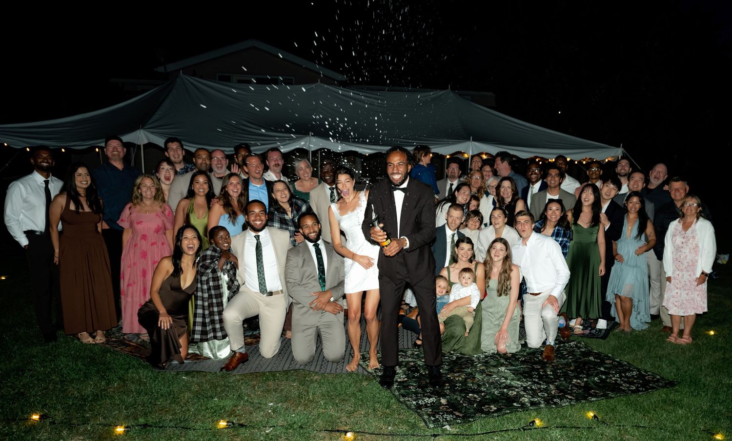Large wedding party group photo at night outside a tent, guests celebrating with confetti.