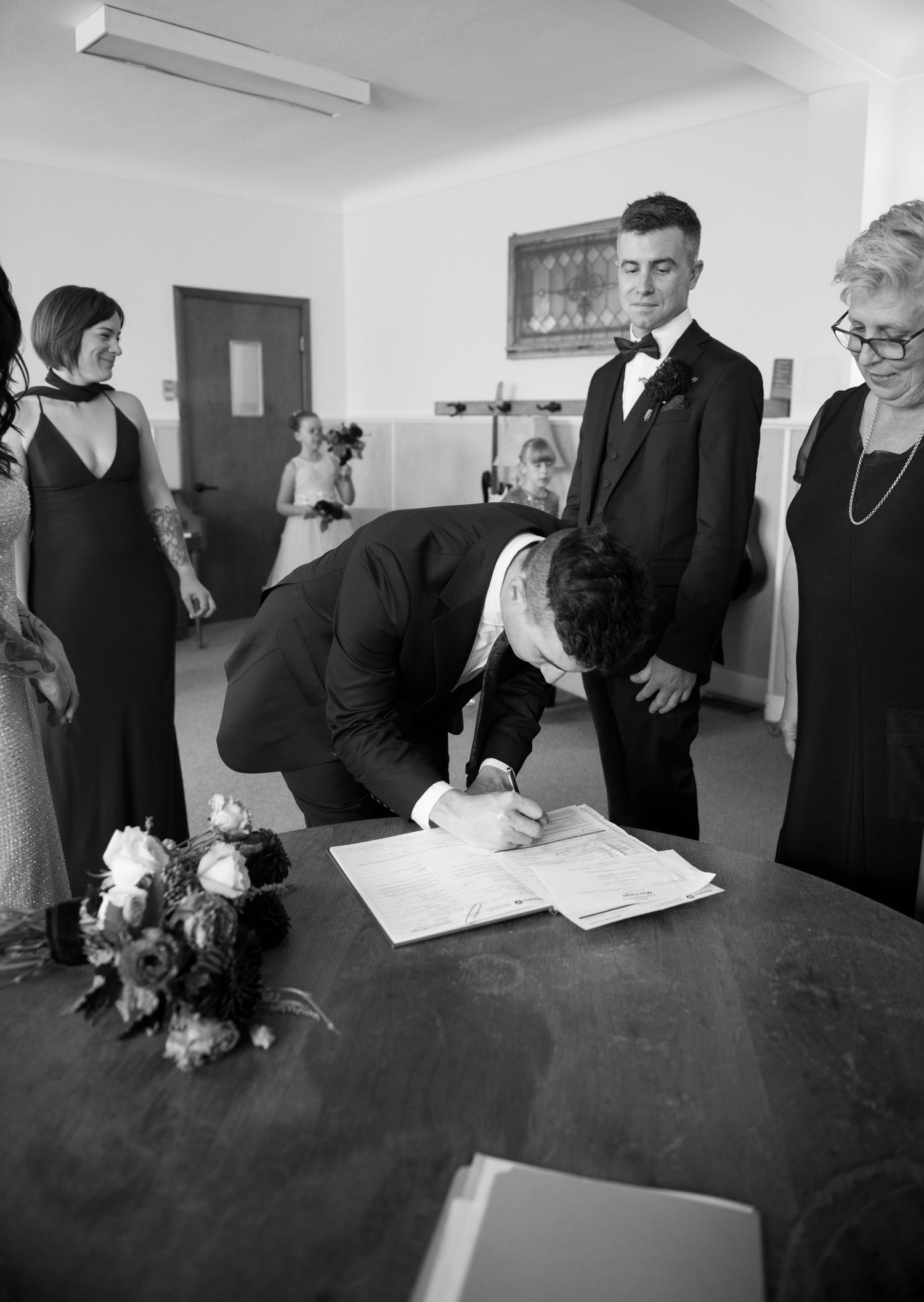 Groom signing wedding register at ceremony table surrounded by family in formal attire, black and white photo.