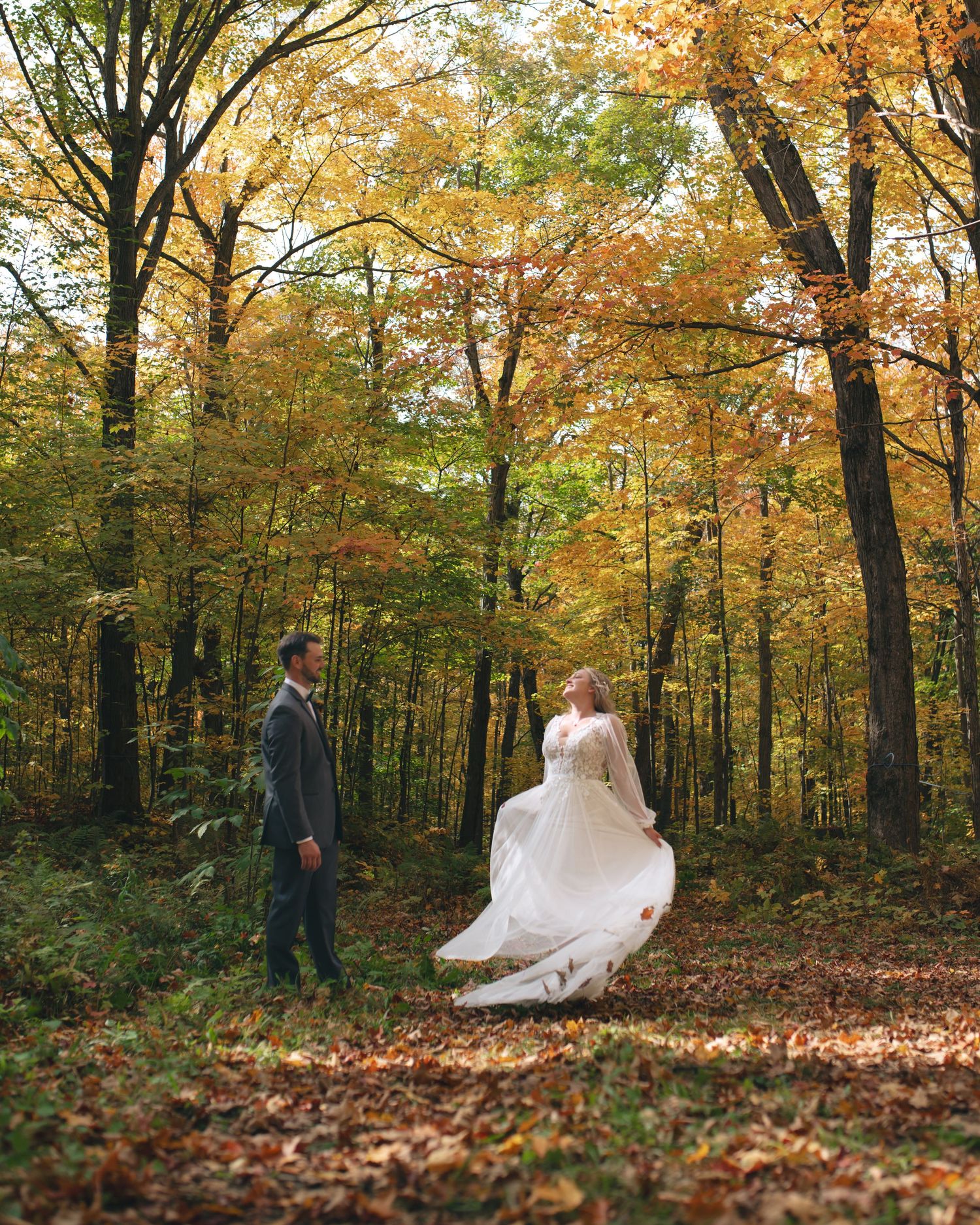 A bride in a flowing white gown twirls with her groom in an autumn forest filled with golden and orange foliage.