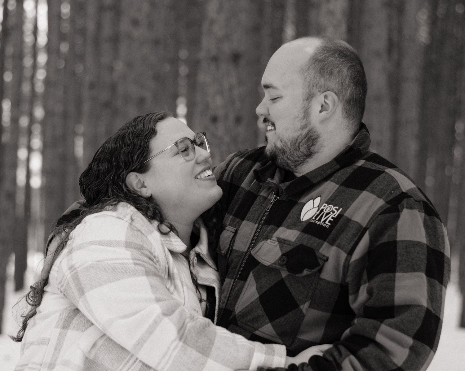 Black and white photo of a smiling couple embracing outdoors in a snowy forest setting.