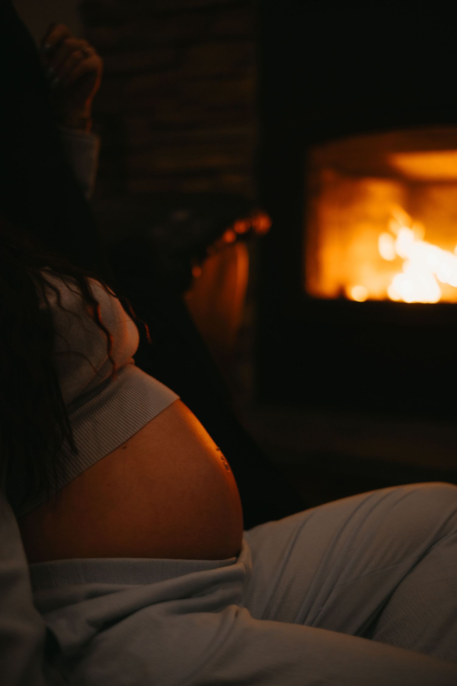Pregnant woman relaxing near a glowing fireplace, warm light illuminating her baby bump from behind.