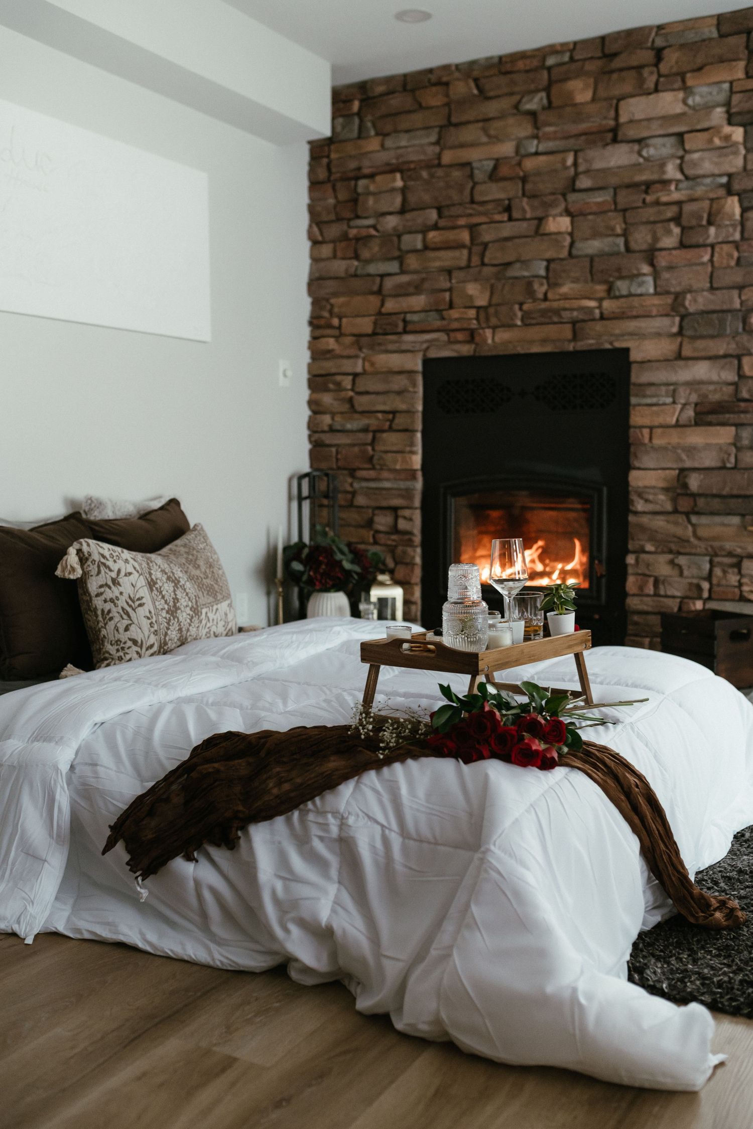 Cozy bedroom with white bedding, breakfast tray, red roses, and a roaring stone fireplace.
