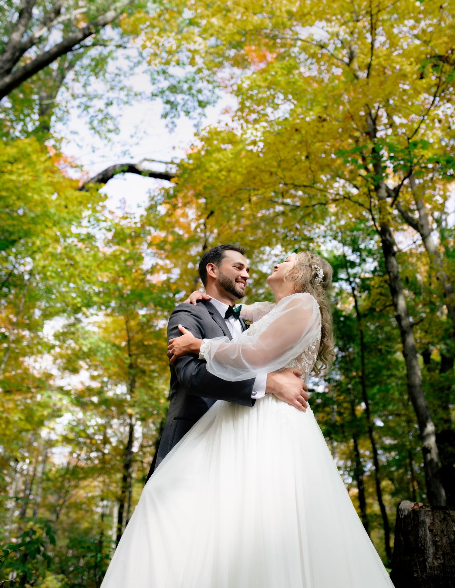A romantic couple in wedding attire dances and embraces among autumn trees with golden yellow foliage.