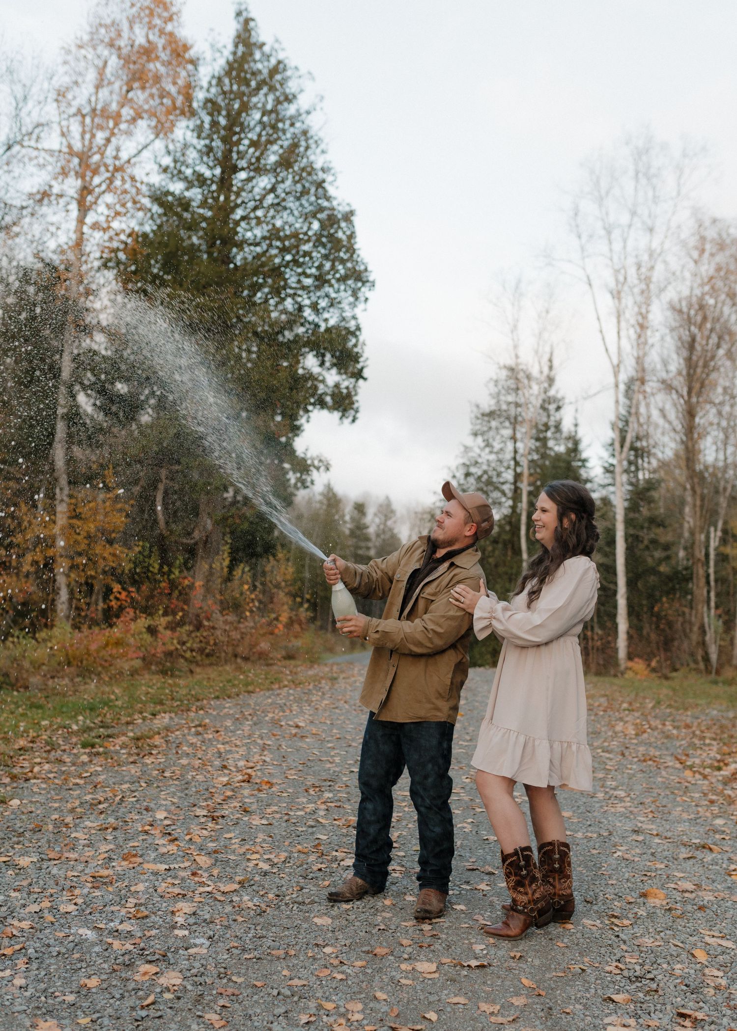 A couple sprays champagne in celebration while standing on fallen autumn leaves in a wooded area.
